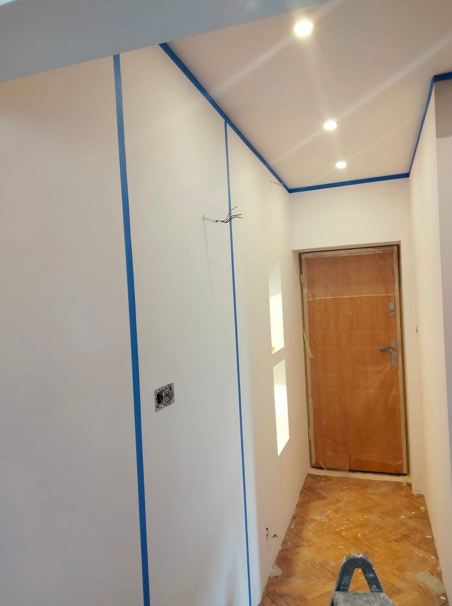 Białe ściany z niebieską taśmą malarską, przygotowane do malowania. Widoczne gniazdka, drewniana podłoga i drzwi zabezpieczone folią. Oświetlenie sufitowe.