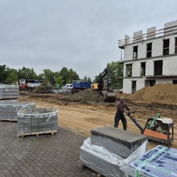 KRET-BRUK SP. Z O.O. - Prace ziemne w Chojnicach: operator zagęszczarki ubija piasek pod kostkę brukową. W tle budynek w stanie surowym i palety z kostką.