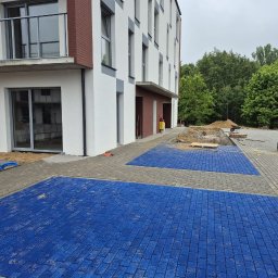 KRET-BRUK SP. Z O.O. - Ułożona, niebieska kostka brukowa na parkingu przed nowoczesnym budynkiem mieszkalnym. Część parkingu wyłożona szarą kostką, w tle widoczny teren przygotowany pod dalsze prace.