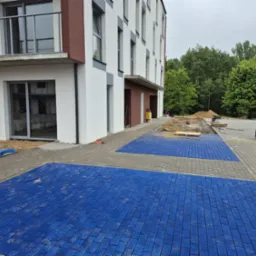 Ułożona, niebieska kostka brukowa na parkingu przed nowoczesnym budynkiem mieszkalnym. Część parkingu wyłożona szarą kostką, w tle widoczny teren przygotowany pod dalsze prace.