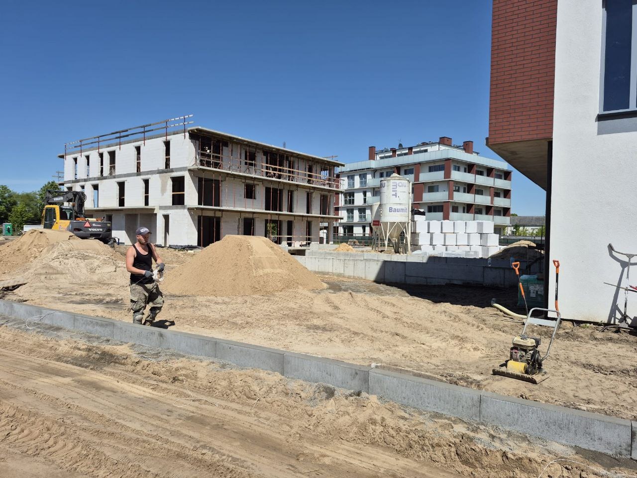 Budowa osiedla: pracownik w terenie z ubijarką, w tle budynki w stanie surowym, góra piasku, betonowe elementy i zbiornik Baumi.