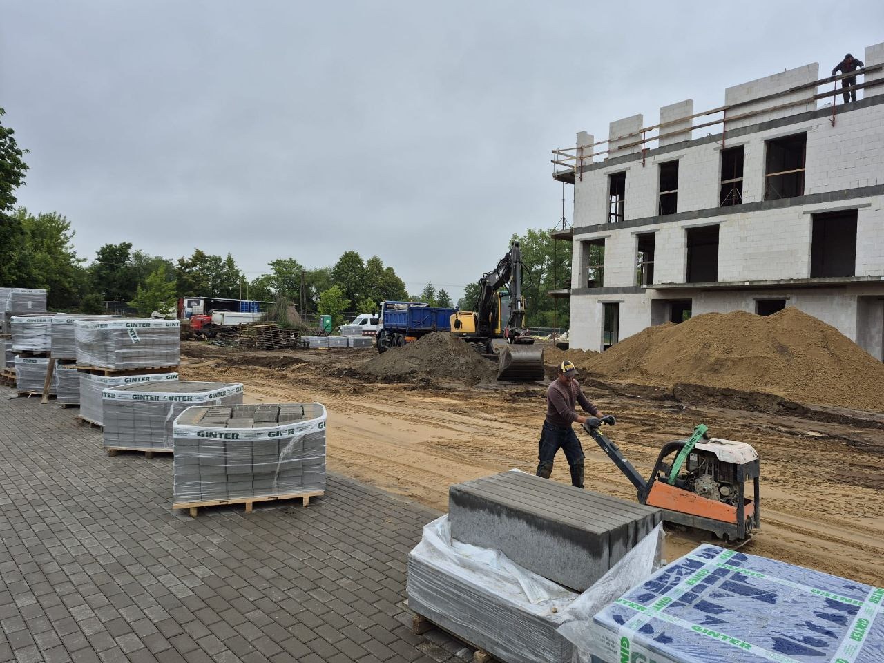 Prace ziemne w Chojnicach: operator zagęszczarki ubija piasek pod kostkę brukową. W tle budynek w stanie surowym i palety z kostką.