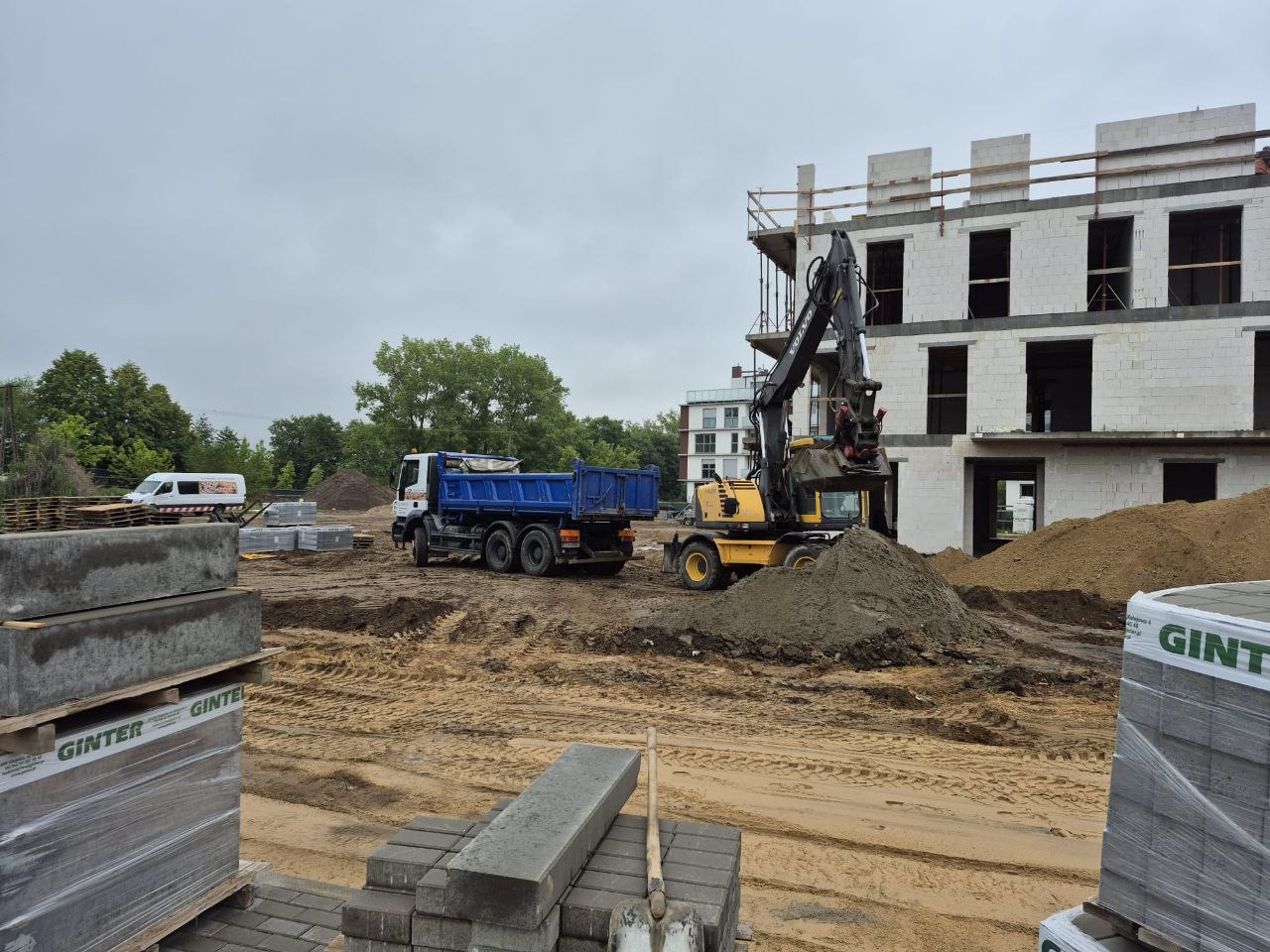 Żółta koparka nabiera ziemię przy budynku w budowie; w tle niebieska ciężarówka, palety z kostką i zaplecze budowy. Pochmurne niebo.