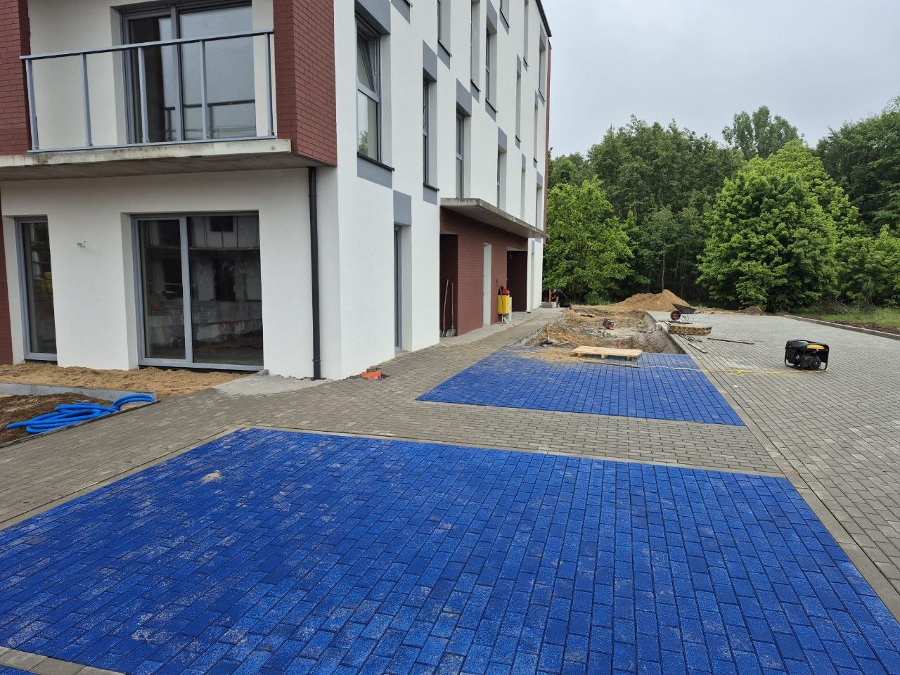 Ułożona, niebieska kostka brukowa na parkingu przed nowoczesnym budynkiem mieszkalnym. Część parkingu wyłożona szarą kostką, w tle widoczny teren przygotowany pod dalsze prace.