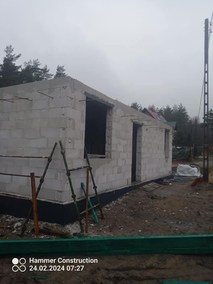 Budowa domu z białych bloczków: ściany w stanie surowym, widoczne otwory okienne i drzwiowe, drabina oparta o ścianę, teren budowy.