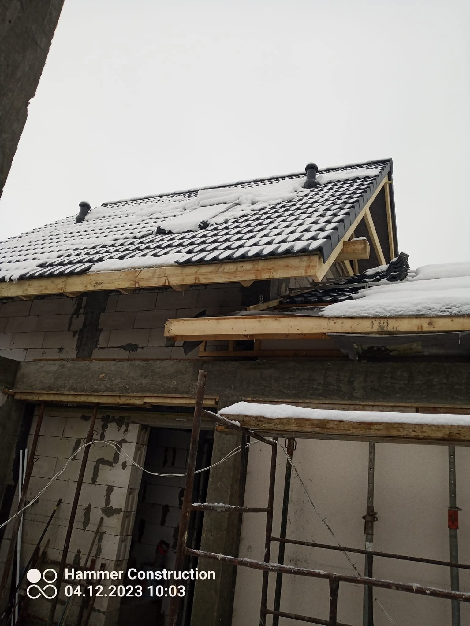 Wznoszenie domu z pustaków: fragment dachu z czarną dachówką, częściowo pokrytą śniegiem, widoczny na tle surowych, niewykończonych ścian i rusztowań.