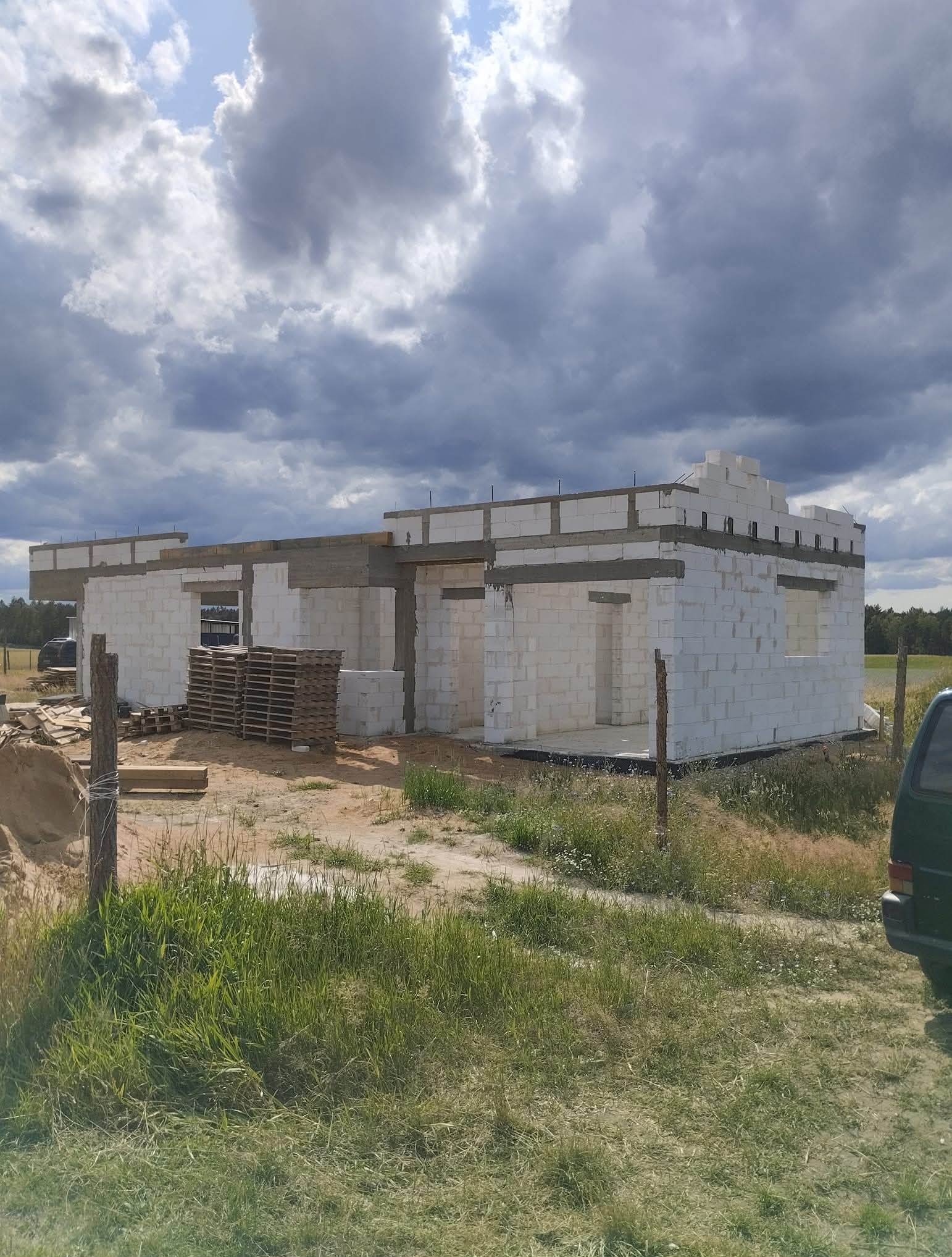 Wznoszenie ścian parterowego domu z białych bloczków, widoczne palety z materiałem budowlanym. W tle pochmurne niebo i zieleń, ogrodzenie z drewnianych słupków.