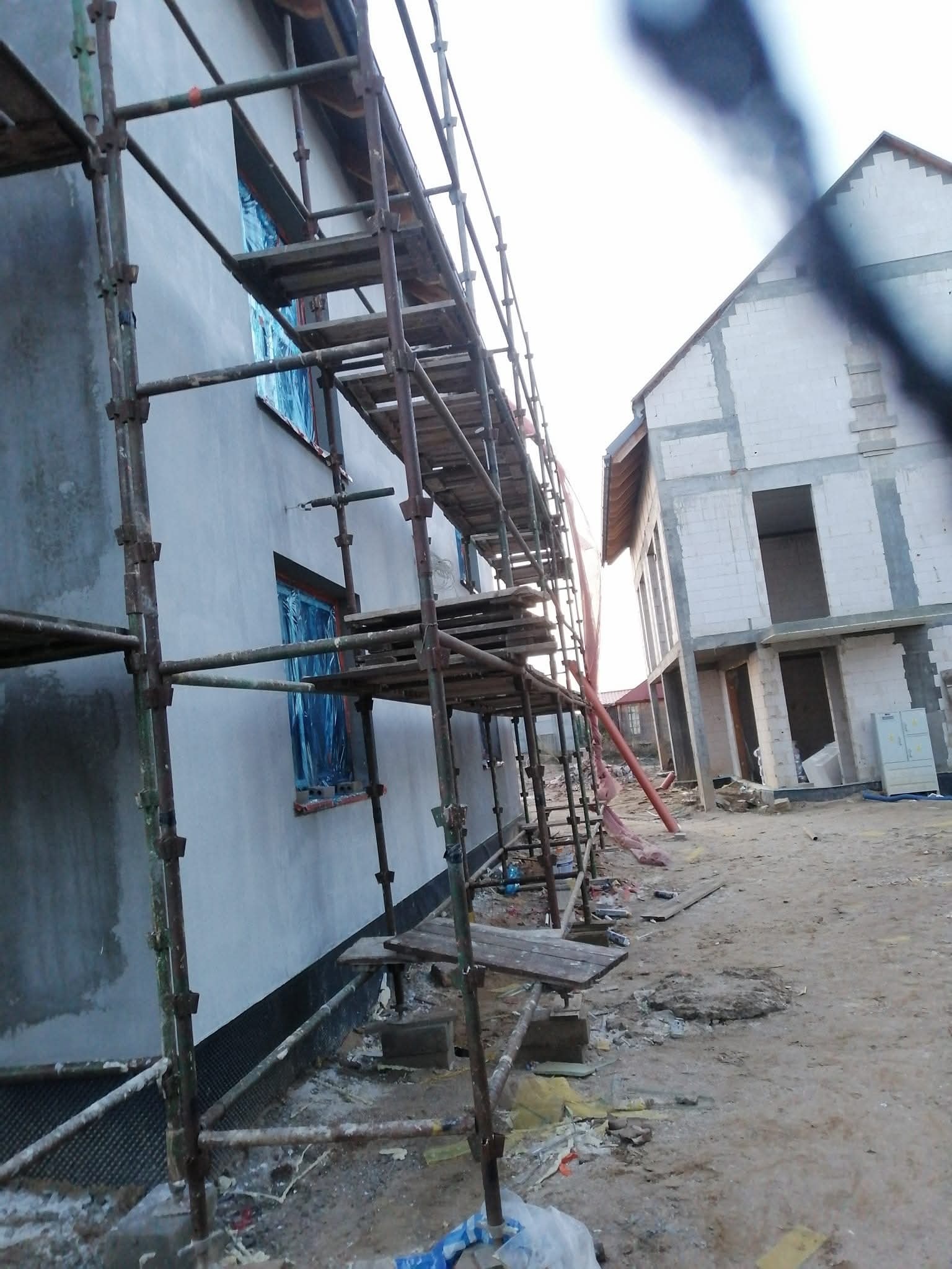 Budowa domu w Dargolezie: ściany otynkowane, rusztowania przy budynku. Okna zabezpieczone folią. Na drugim planie budynek w trakcie murowania.