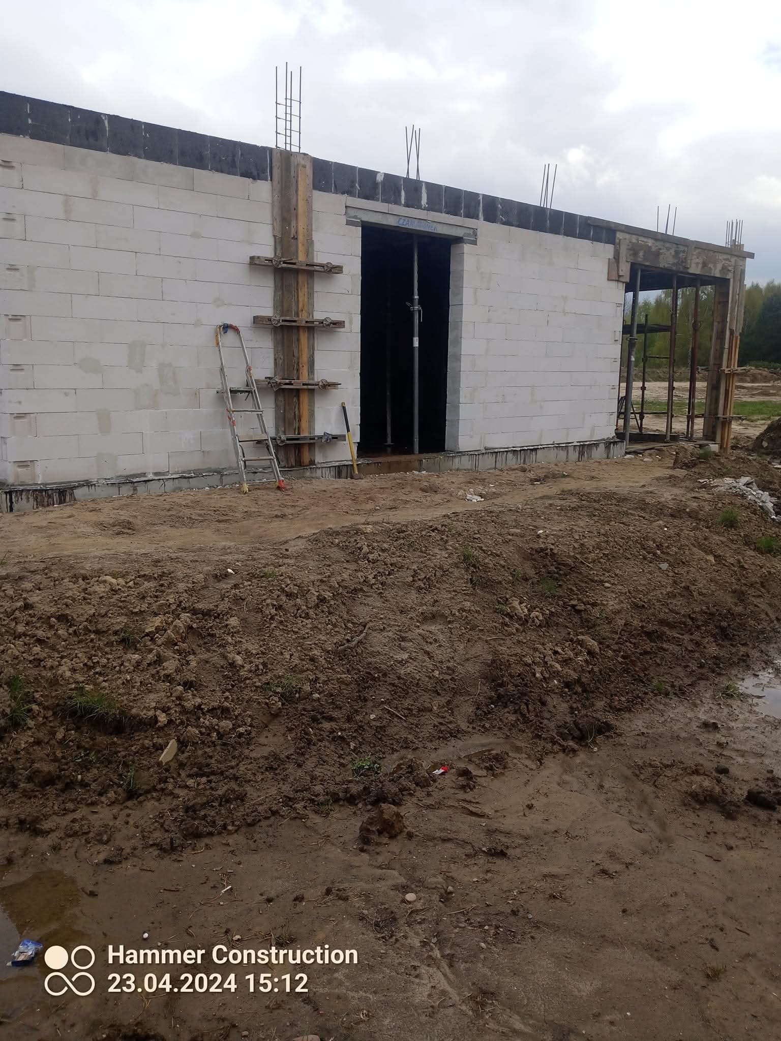 Ściana budynku w trakcie wznoszenia z białych bloczków, widoczne zbrojenia i szalunki. Na pierwszym planie ziemia i błoto, w tle drzewa.