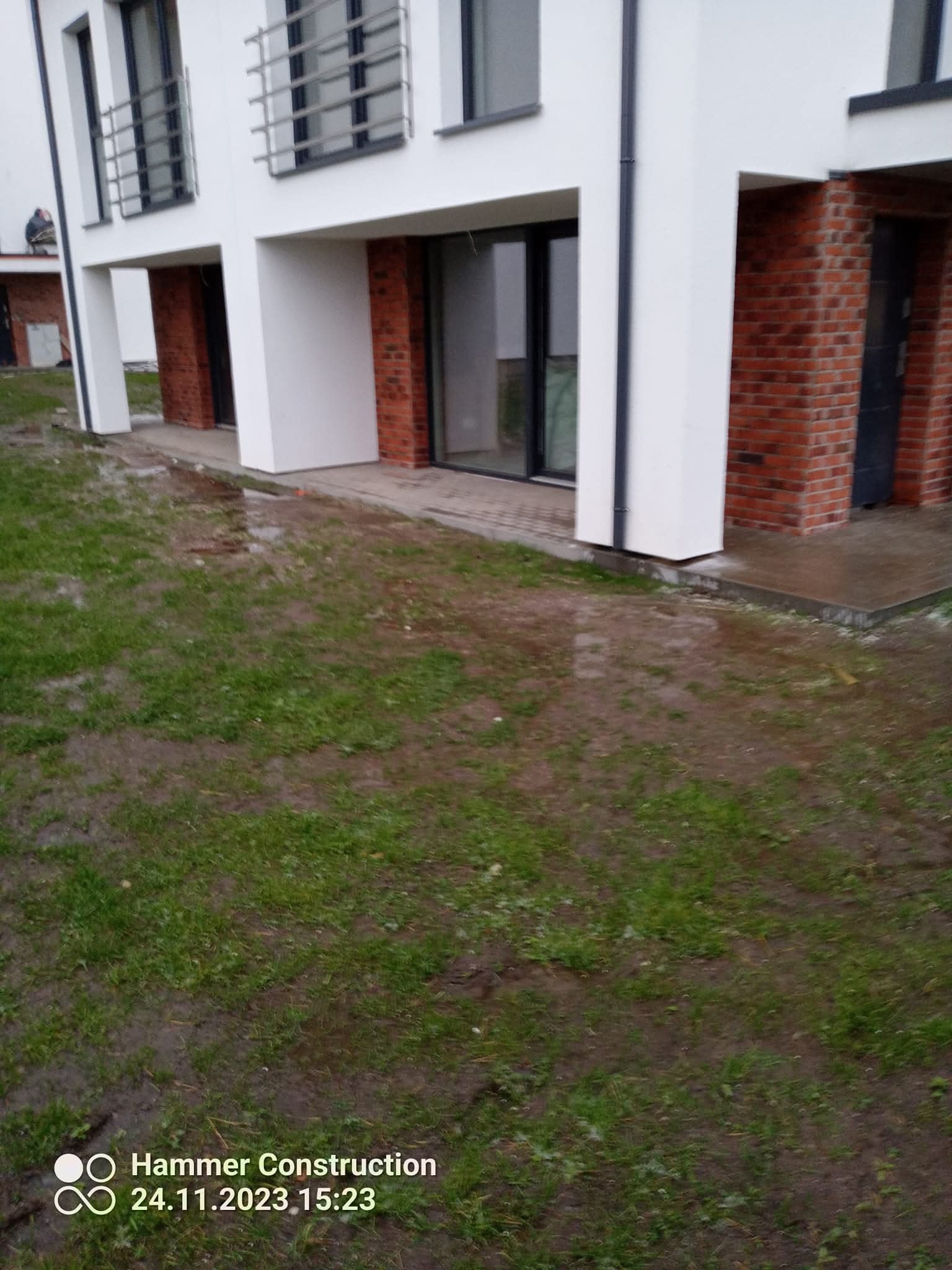 Nowoczesny, dwupiętrowy dom z białymi ścianami i ceglanymi elementami wykończeniowymi, widziany z perspektywy trawnika w pochmurny dzień. Okna i drzwi w ciemnych ramach.