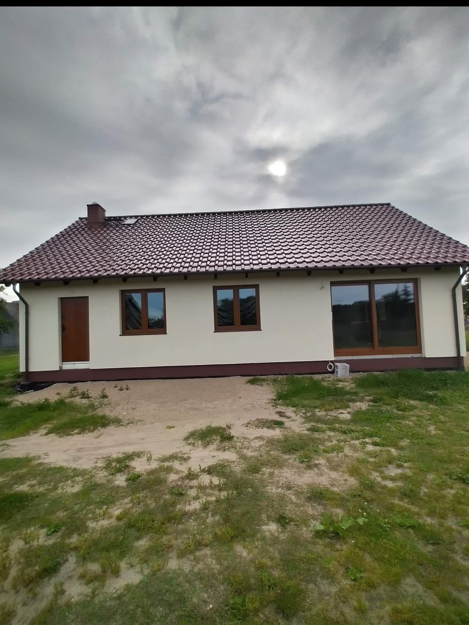 Parterowy dom z brązowym dachem, brązowymi oknami i drzwiami na tle pochmurnego nieba. Widoczne elementy wykończenia elewacji i otoczenia budynku.