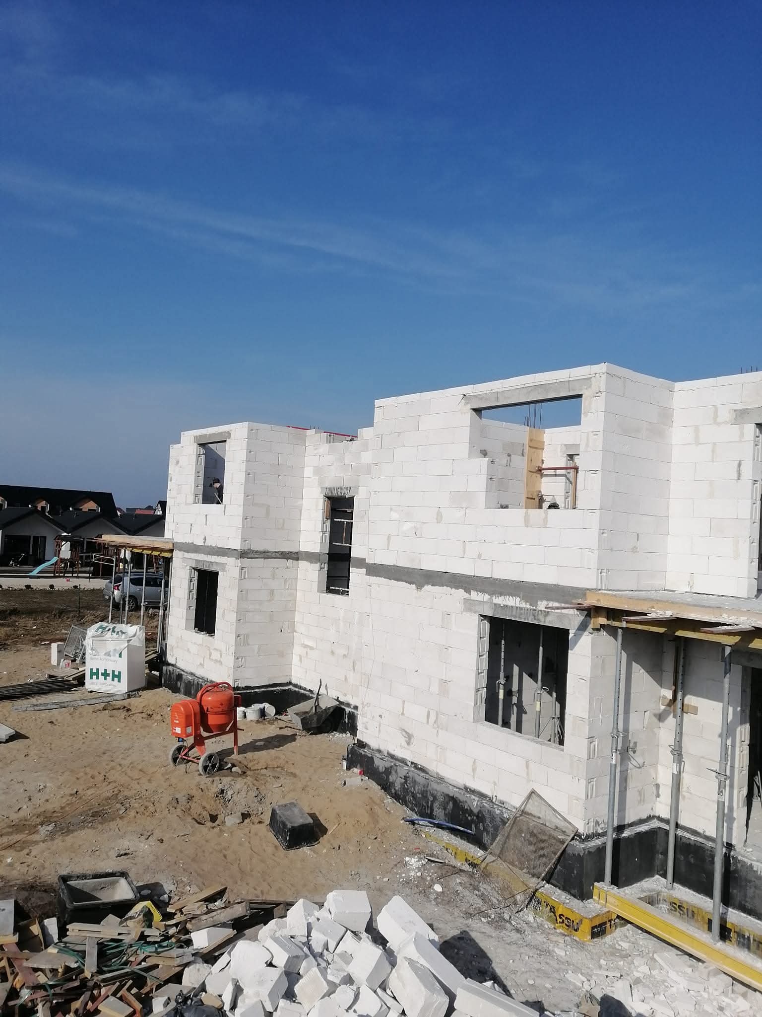 Wznoszenie ścian z białych bloczków na budowie domu. Widoczna betoniarka, materiały budowlane i fragmenty konstrukcji. Błękitne niebo w tle.