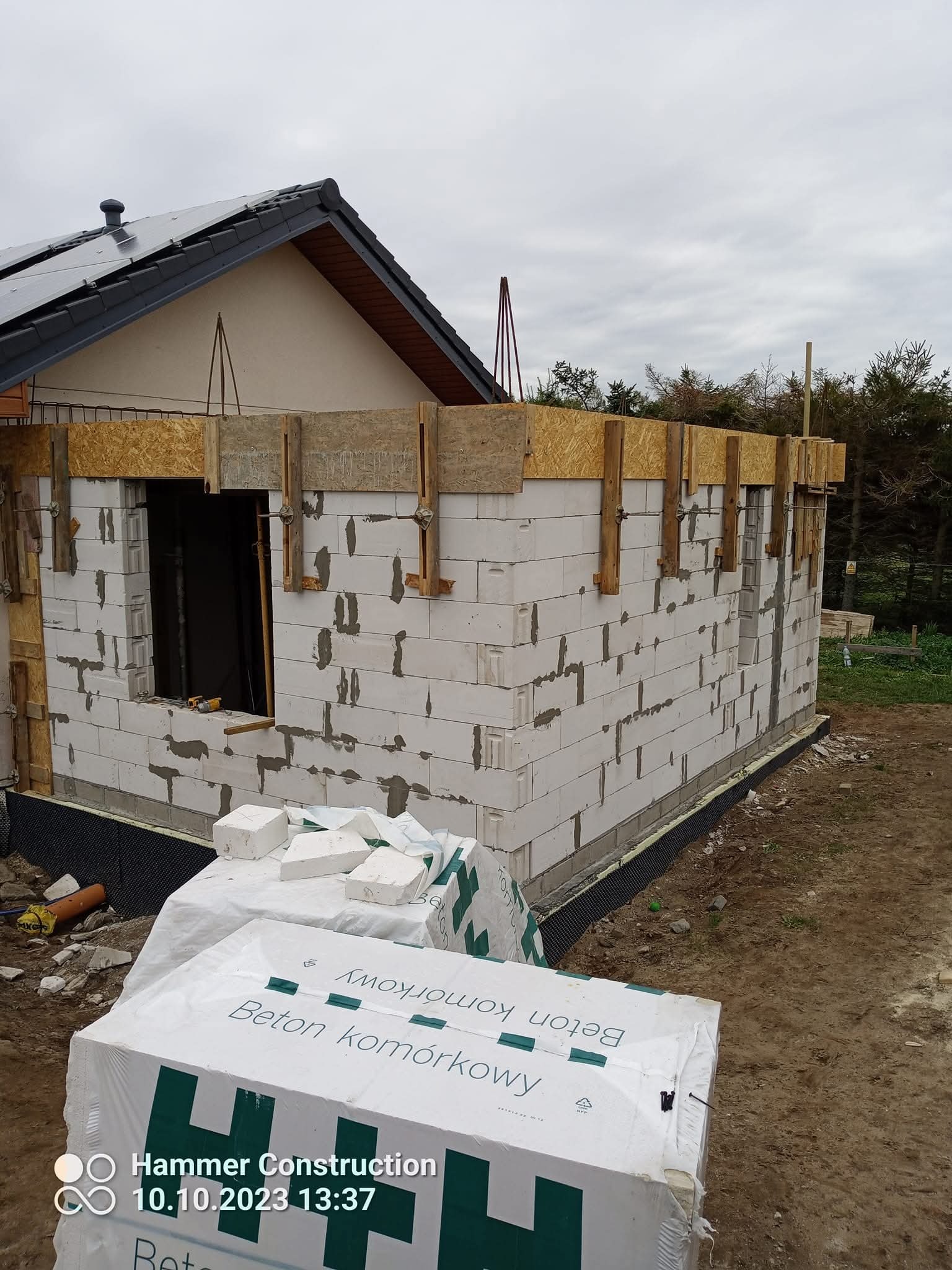 Budowa domu z betonu komórkowego: ściany wzniesione do poziomu okien, widoczne szalunki i zapakowane bloczki. Dach z panelami solarnymi w tle.