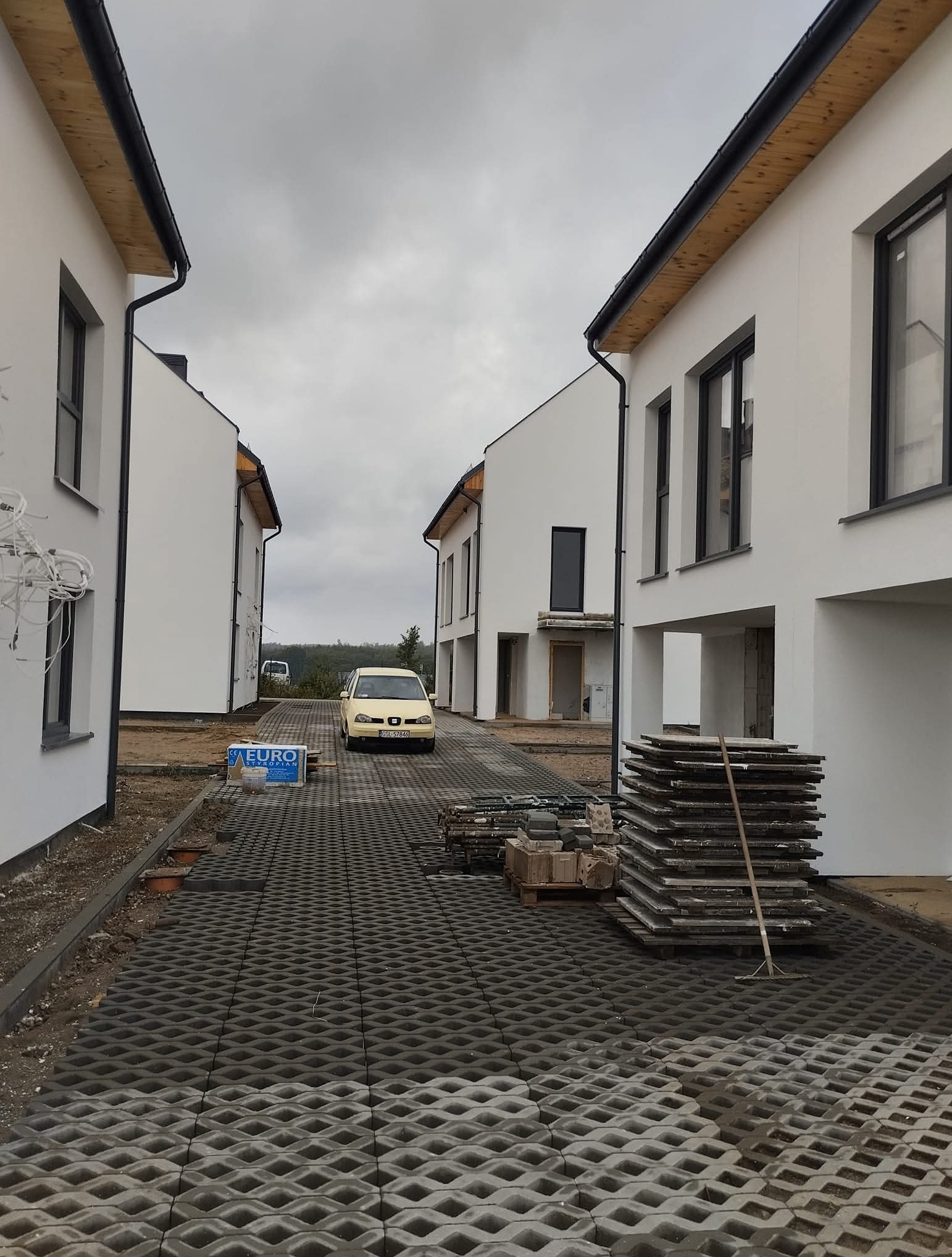 Osiedle w trakcie budowy: białe domy szeregowe, ułożona ażurowa kostka brukowa, zaparkowany samochód. Widoczne materiały budowlane i narzędzia.