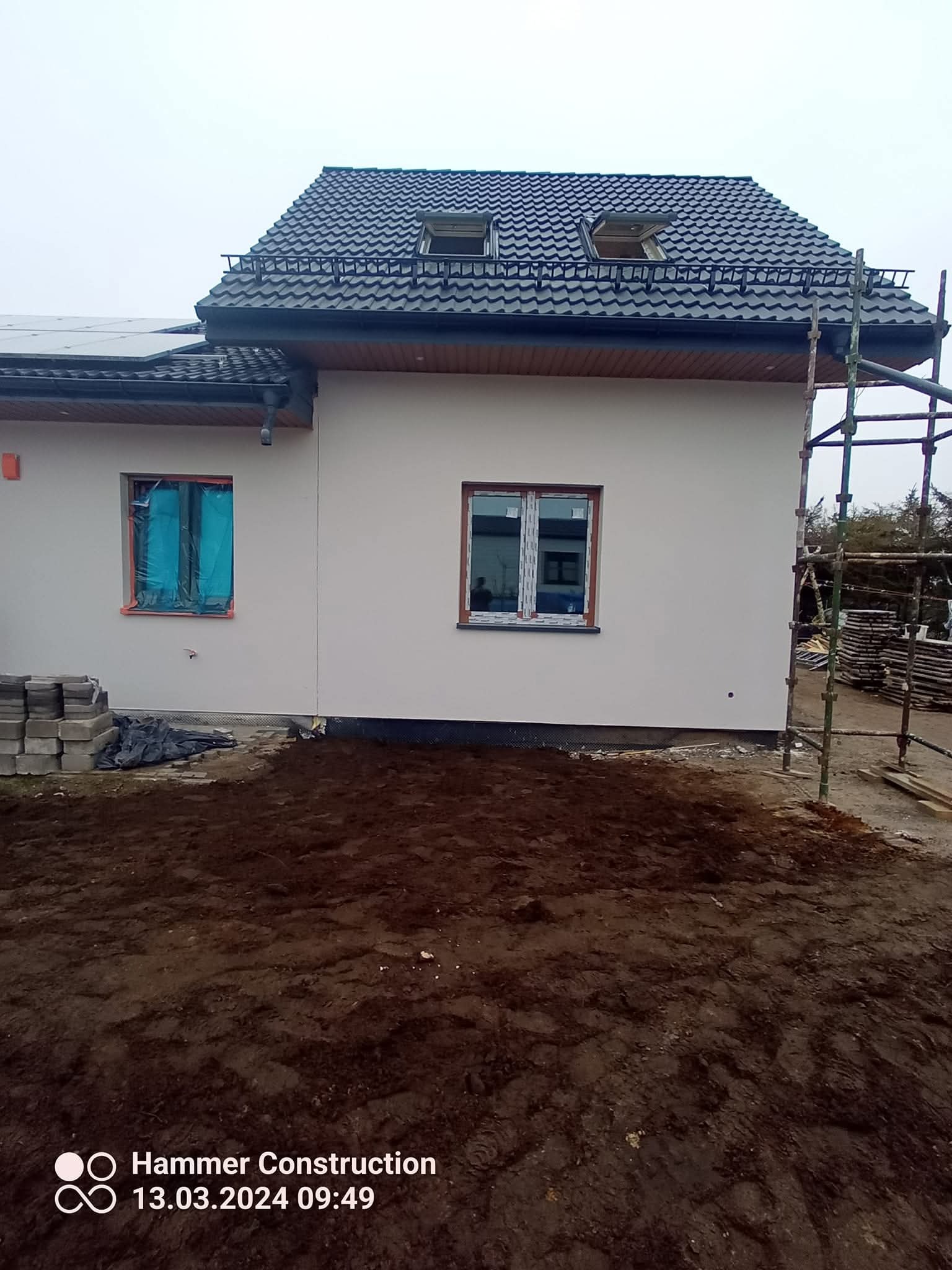 Boczny widok domu z oknami zabezpieczonymi folią, rusztowaniem i stertą kostki brukowej. Widoczne panele słoneczne i dach z oknami.