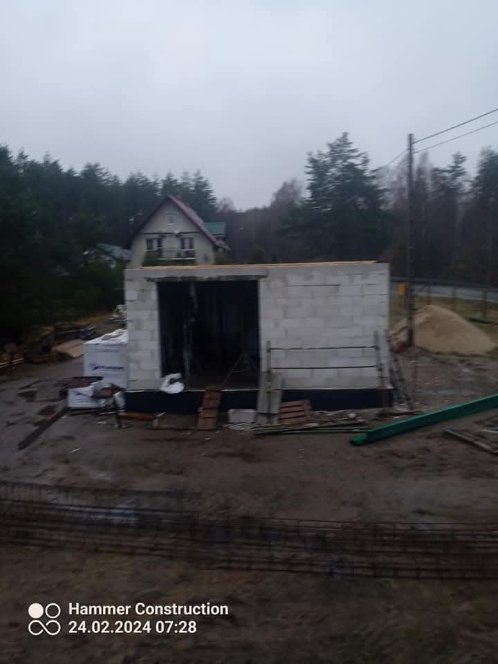 Wznoszenie ścian parterowego budynku z bloczków betonowych na fundamencie. Widoczne zbrojenie i elementy konstrukcyjne. W tle dom i zalesiony teren.