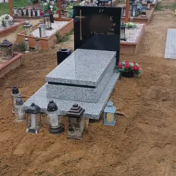 Granitowy nagrobek z czarną tablicą i złotym krzyżem na cmentarzu, otoczony lampionami i świeżo usypaną ziemią. W tle inne groby i drzewa.