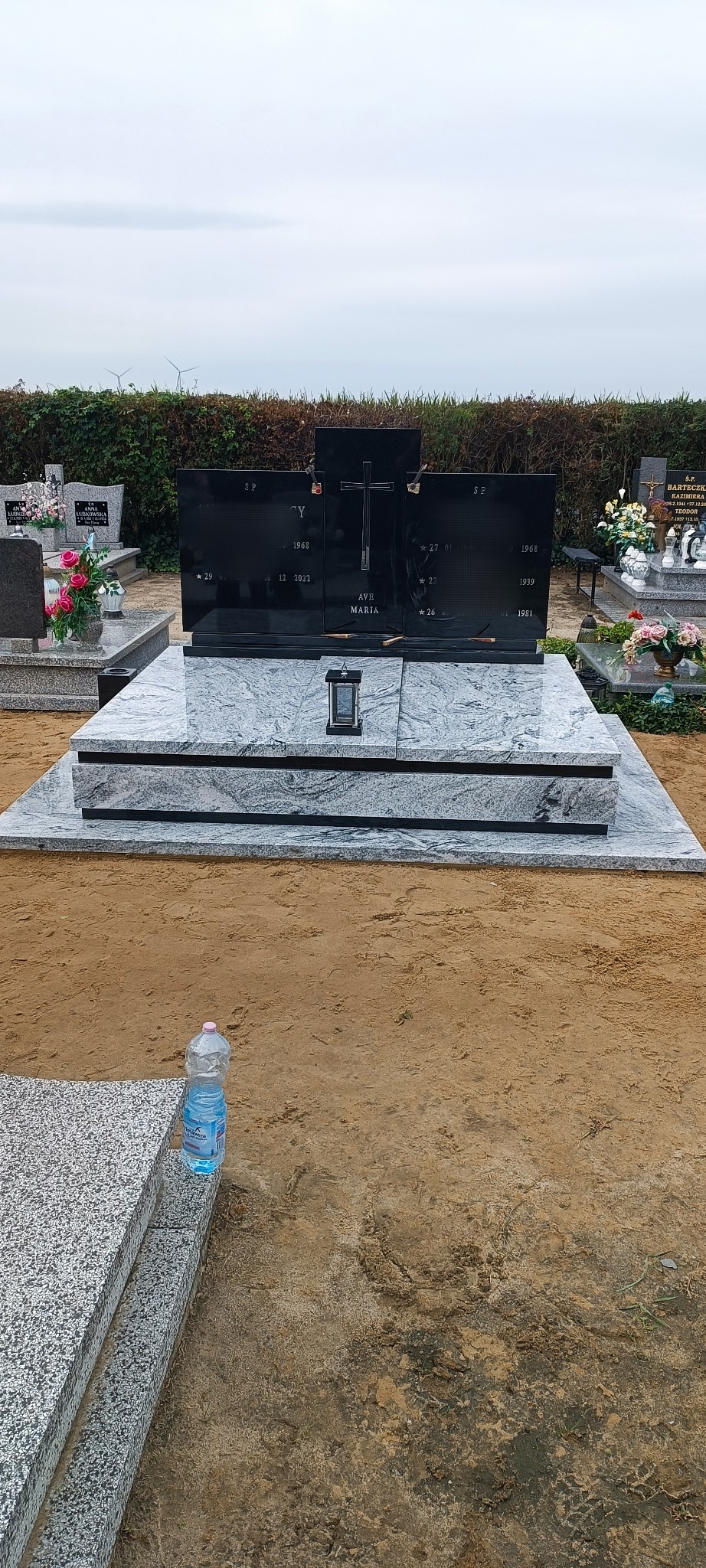 Czarny, podwójny nagrobek z krzyżem na cmentarzu, otoczony innymi nagrobkami. Na pierwszym planie butelka wody. Tło: zielone krzewy i pochmurne niebo.