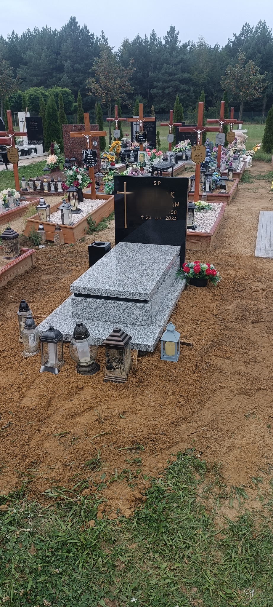 Granitowy nagrobek z czarną tablicą i złotym krzyżem na cmentarzu, otoczony lampionami i świeżo usypaną ziemią. W tle inne groby i drzewa.
