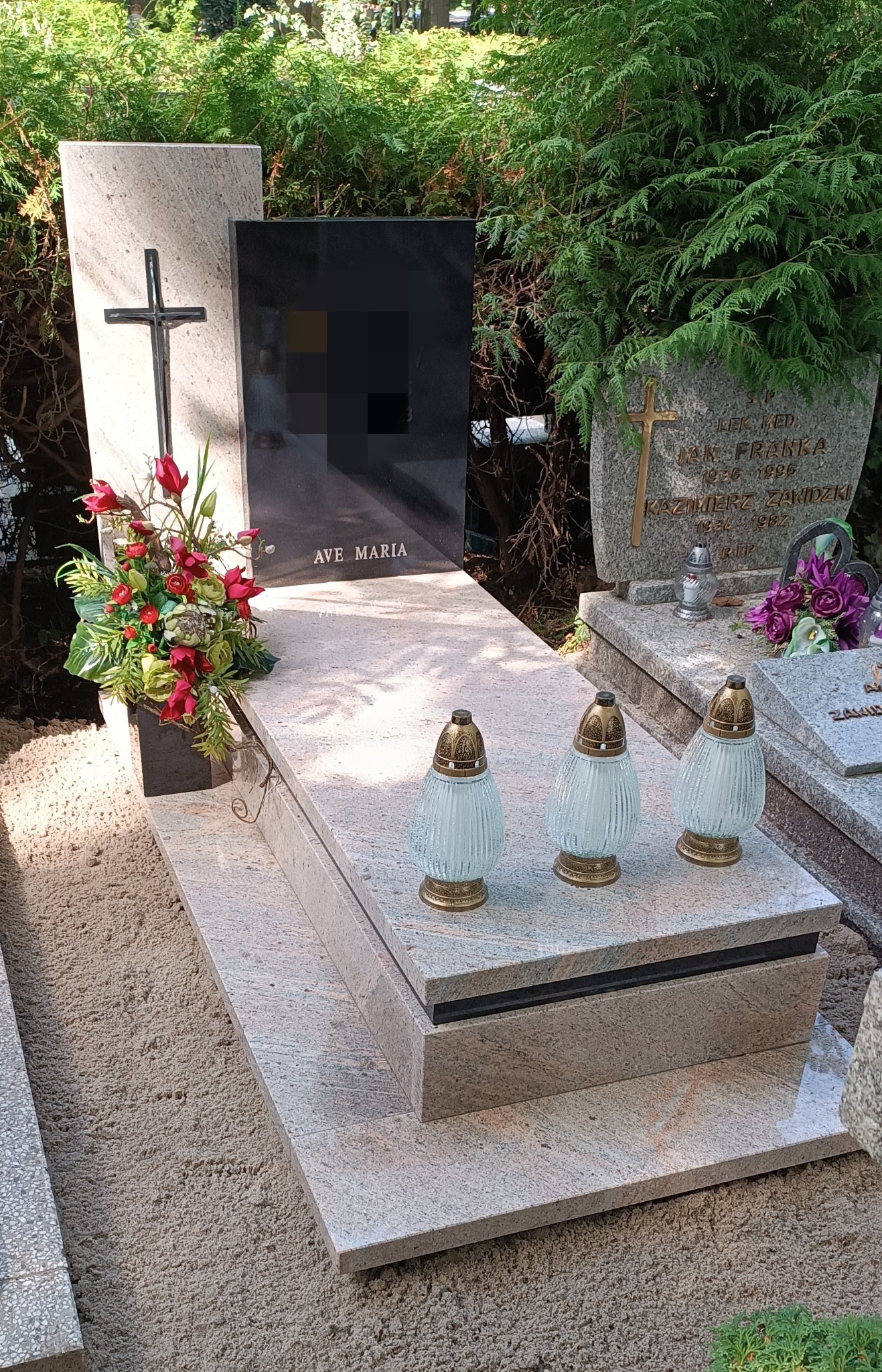 Jasny, granitowy nagrobek z czarną tablicą i napisem 'Ave Maria', ozdobiony kwiatami i lampionami. W tle zieleń cmentarza.
