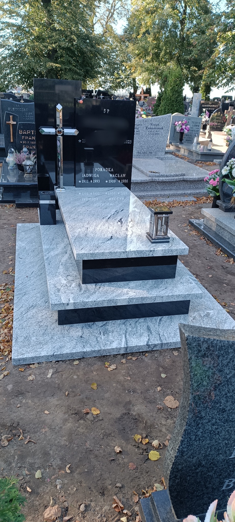 Czarno-szary nagrobek z krzyżem na cmentarzu, z inskrypcjami. Widoczne otoczenie cmentarza, drzewa i inne nagrobki. Jesienne liście na ziemi.