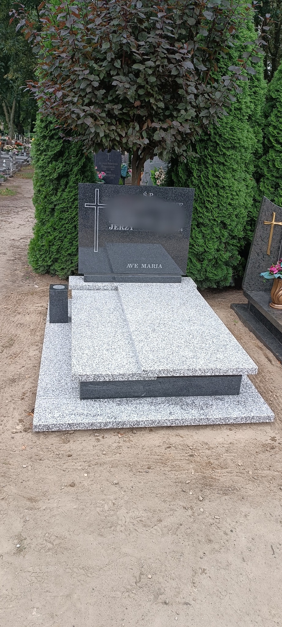 Granitowy nagrobek z krzyżem i napisem 'Ave Maria' na cmentarzu, otoczony zielenią. Pomnik w odcieniach szarości, z widocznym ziarnem kamienia.