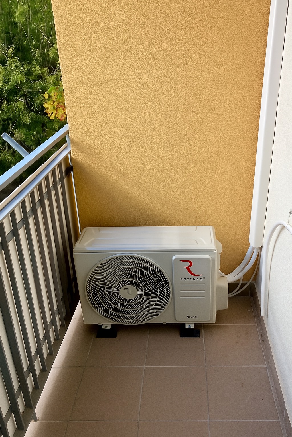 Biała jednostka zewnętrzna klimatyzacji marki Sotenso, zamontowana na balkonie z beżowymi płytkami i żółtą ścianą. Widoczne przewody i fragment balustrady.