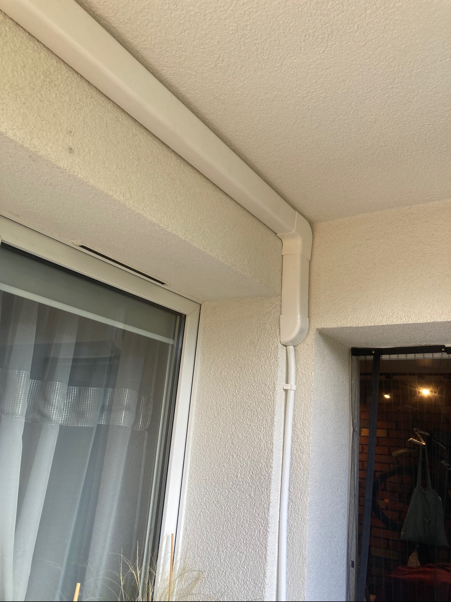 Białe rury klimatyzacji poprowadzone wzdłuż ściany i sufitu, widoczne okno z firanką i fragment balkonu/tarasu.