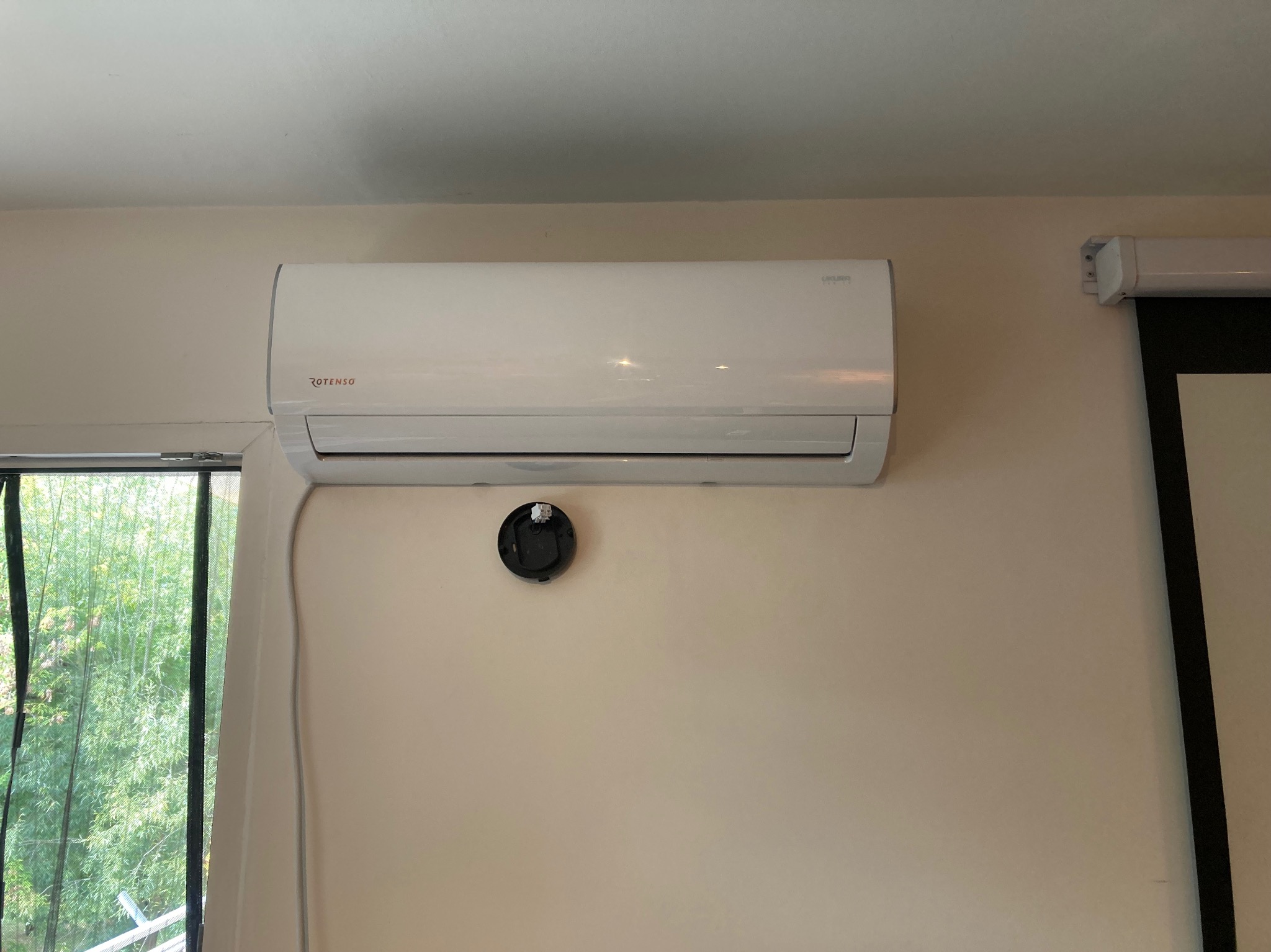Biała klimatyzacja Rotenso zamontowana na ścianie, obok okna z widokiem na zieleń i zwinięty ekran projekcyjny. Widoczny czujnik na ścianie.