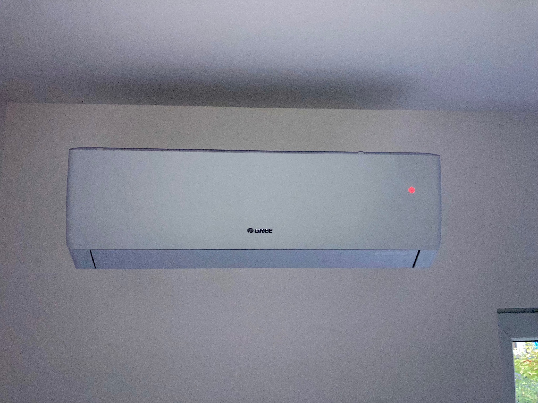 Biała klimatyzacja Gree na ścianie, widoczna czerwona dioda sygnalizacyjna. Fragment okna z zielenią w prawym dolnym rogu kadru.