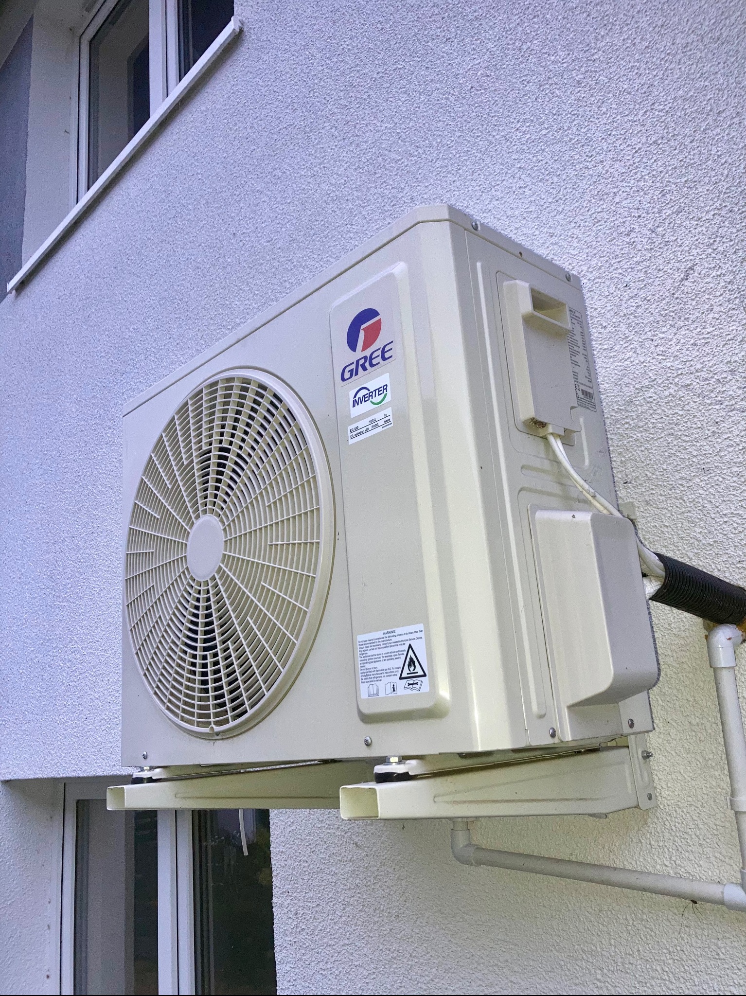 Klimatyzator GREE zamontowany na elewacji budynku, widoczny z boku, z rurami odprowadzającymi. Montaż nad oknem. Teksturowana ściana zewnętrzna.