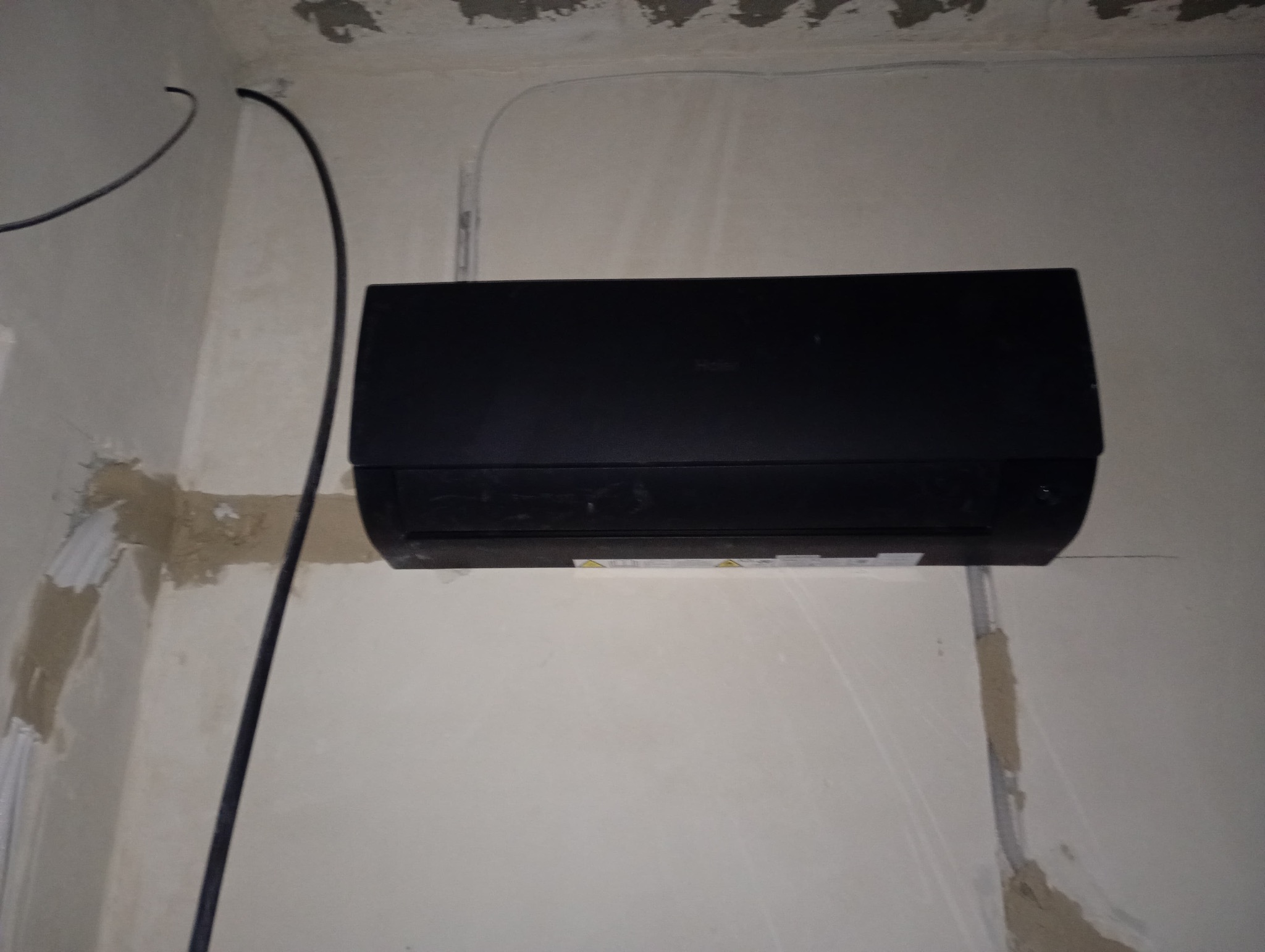 Czarny klimatyzator Haier zamontowany na surowej ścianie z widocznymi przewodami i śladami po instalacji. Widoczne niedoskonałości wykończenia.