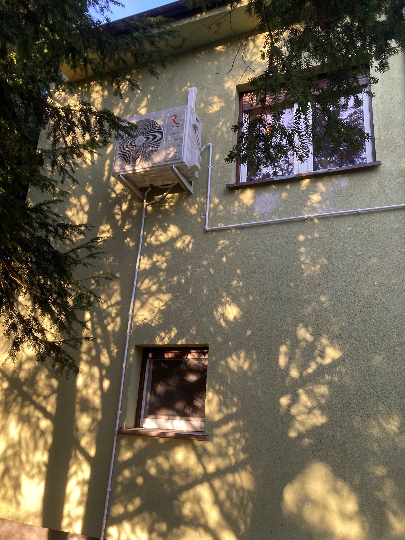 Zamontowany klimatyzator Rotenso na elewacji budynku, widoczne okna i cień drzewa. Biała rurka odprowadzająca skropliny. Ujęcie z dołu, w słoneczny dzień.