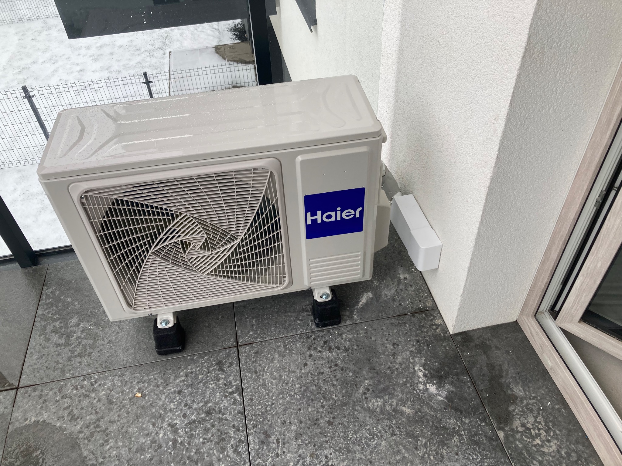 Biała jednostka zewnętrzna klimatyzacji Haier na szarym tarasie, z widocznymi kroplami wody i śniegiem w tle, zamontowana na czarnych podkładkach antywibracyjnych.