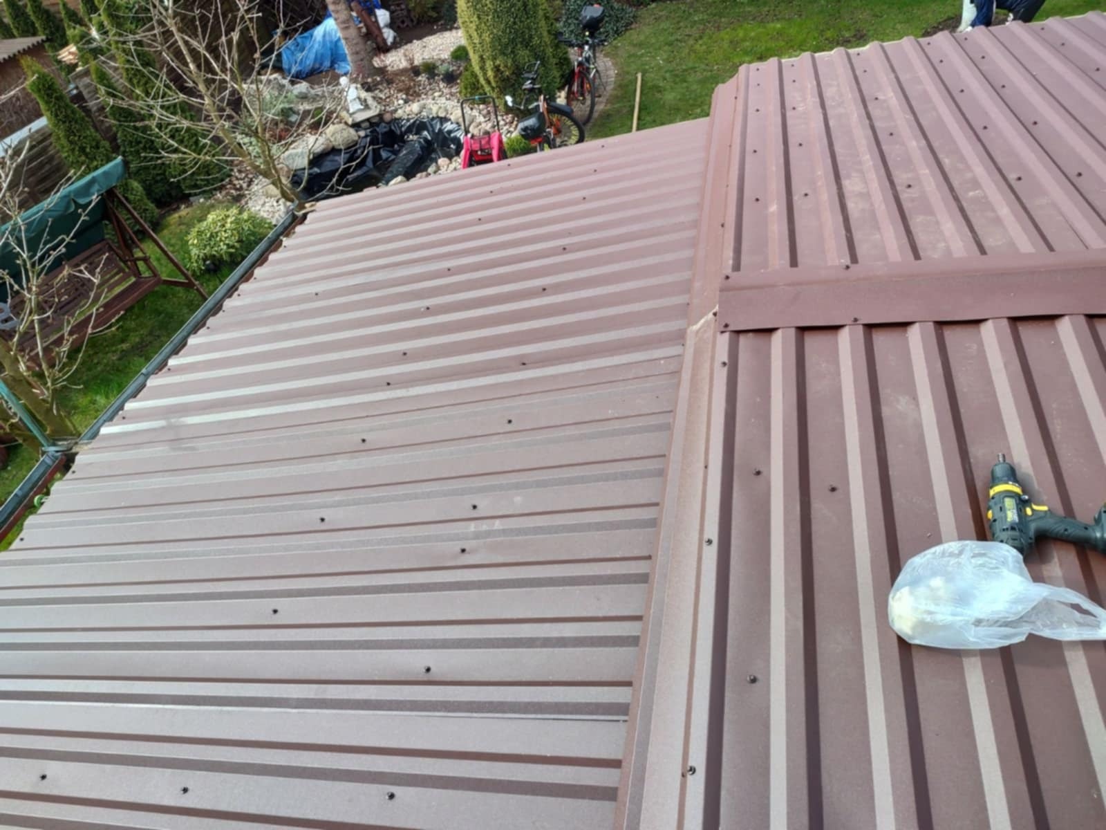 Ukończone krycie dachu blachą trapezową w kolorze brązowym, widoczne wkręty, fragment ogrodu w tle, narzędzia pracy dekarskiej.