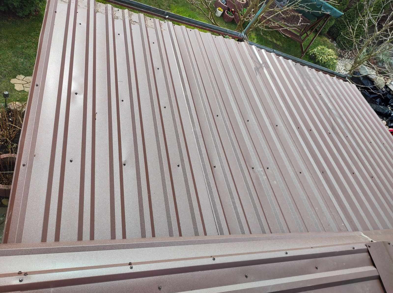 Nowe brązowe krycie dachu z blachy trapezowej, widok z góry. Widoczne mocowania i fragment ogrodu w tle. Blacha ułożona w pionowe pasy.