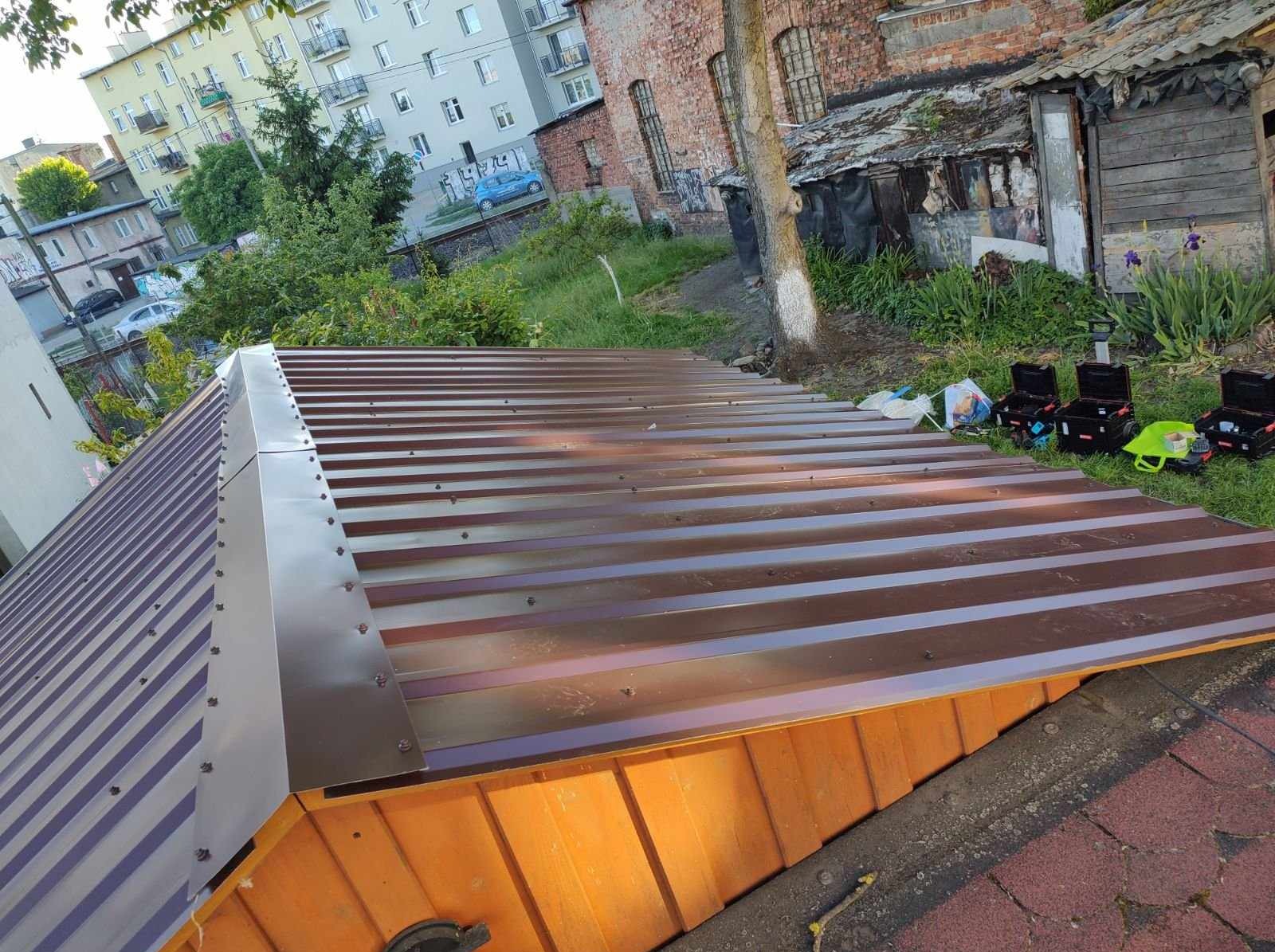 Nowe, brązowe krycie dachowe z blachy trapezowej, zamontowane na drewnianej konstrukcji. W tle budynki mieszkalne i narzędzia dekarskie.