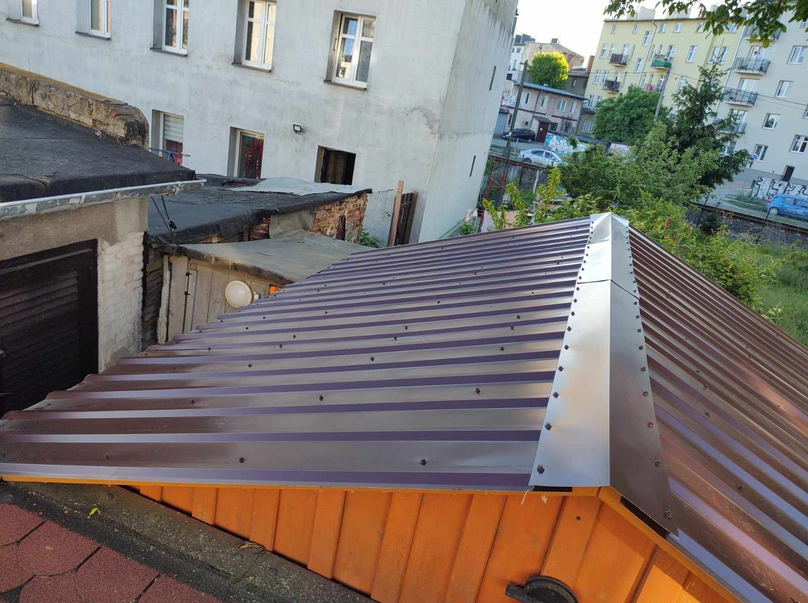 Nowy, brązowy dach z blachy trapezowej z widocznymi śrubami i obróbką blacharską na tle budynków mieszkalnych i zieleni. Ujęcie z góry.