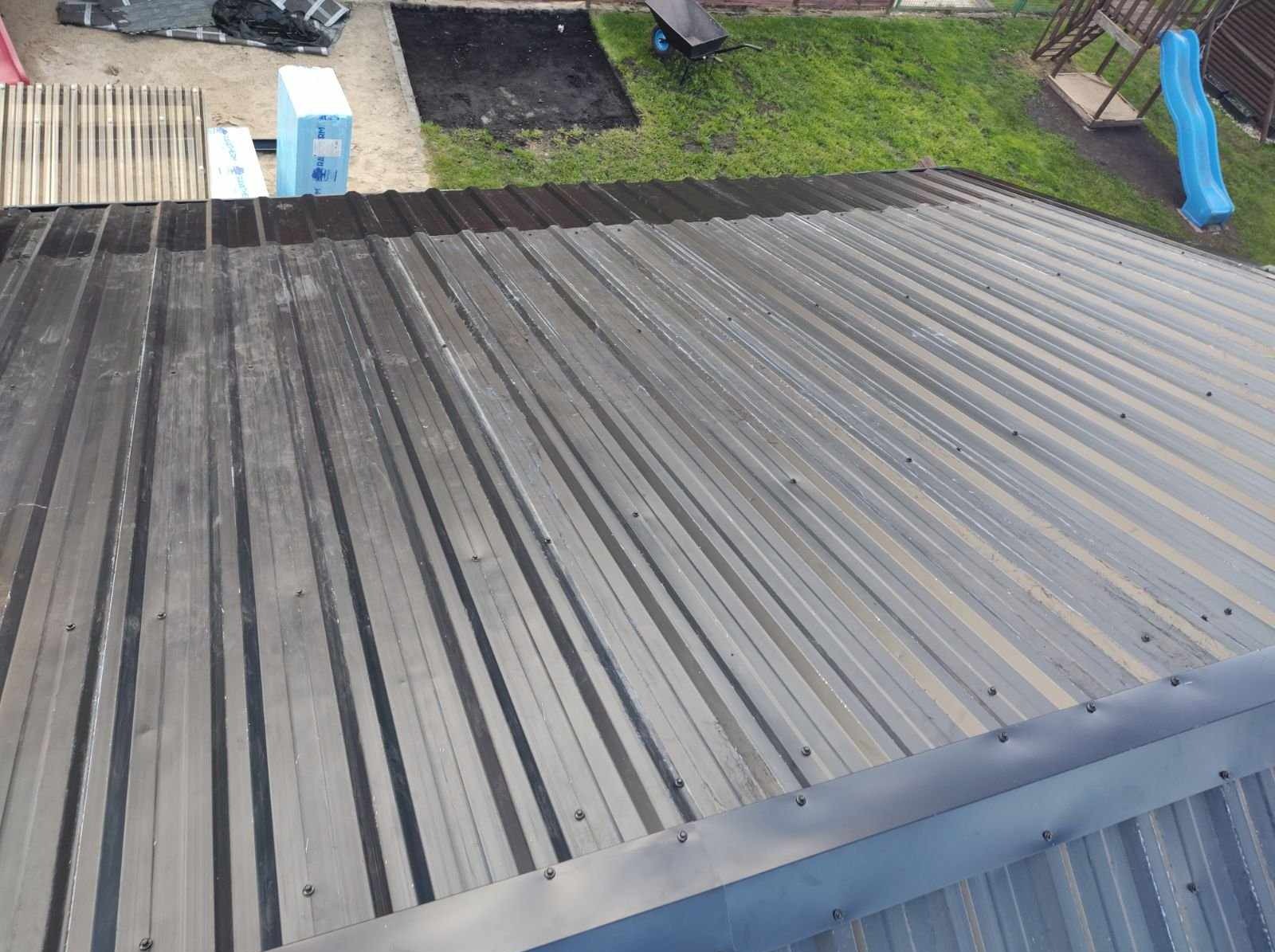 Metalowy dach z widocznymi śladami czyszczenia, częściowo mokry, z bliska. W tle elementy ogrodu i materiały budowlane.