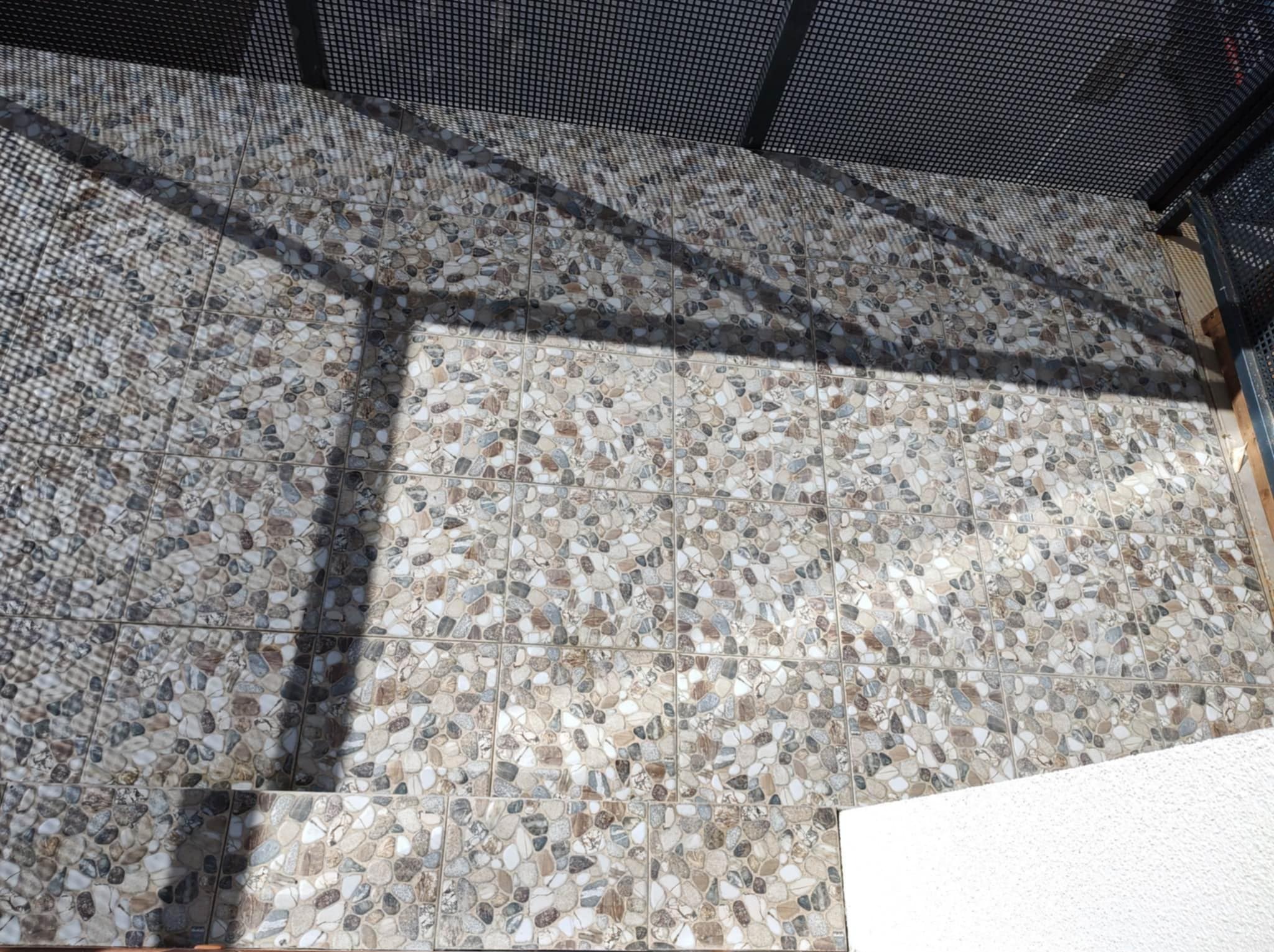 Ułożona podłoga z płytek imitujących kamień na balkonie, z widocznymi cieniami metalowej konstrukcji nad nią. Jasne, naturalne światło podkreśla teksturę płytek.