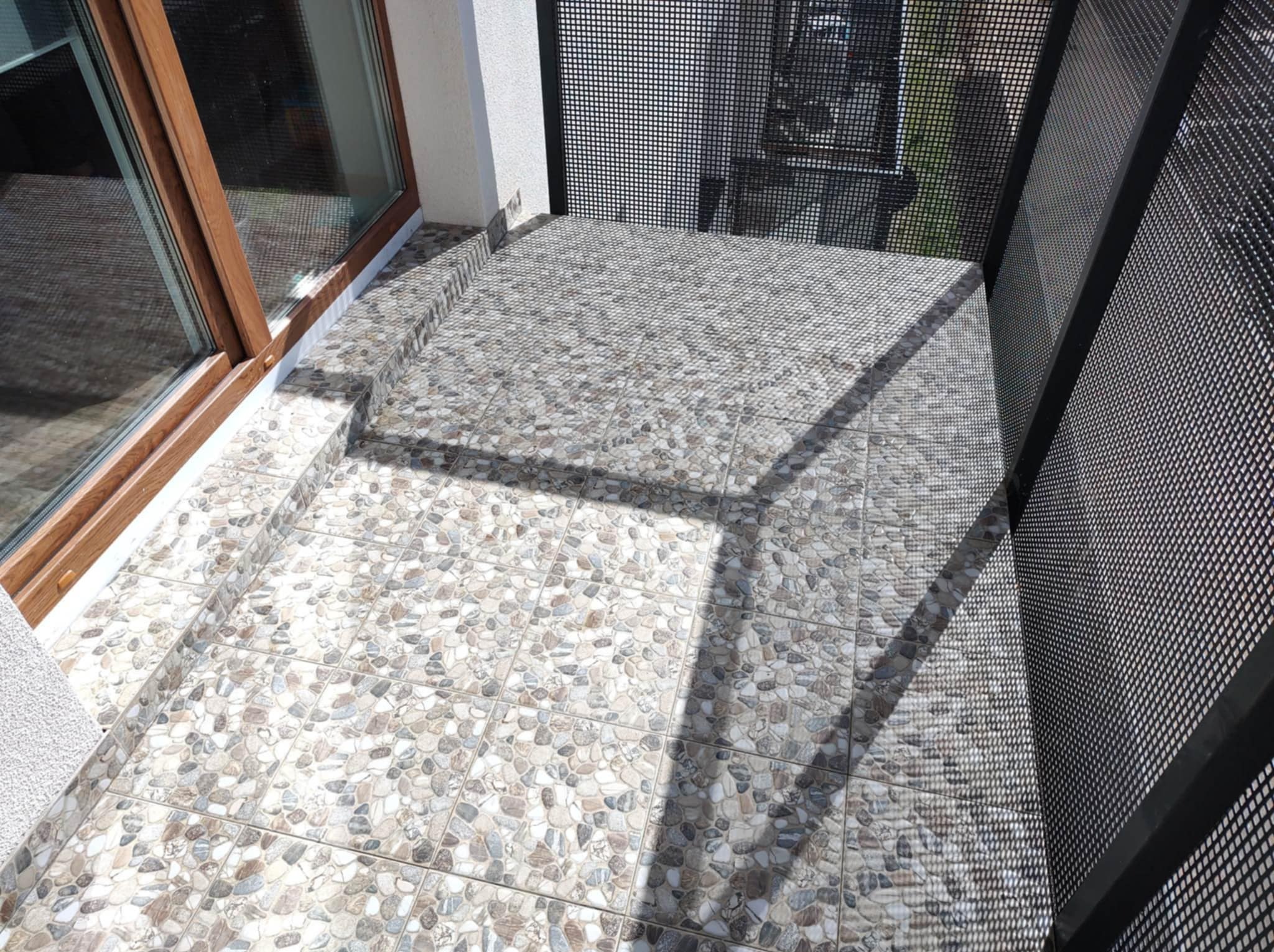 Wykończony balkon z szarymi płytkami imitującymi kamień, czarna metalowa balustrada z siatki, brązowe okno balkonowe. Widok z góry.