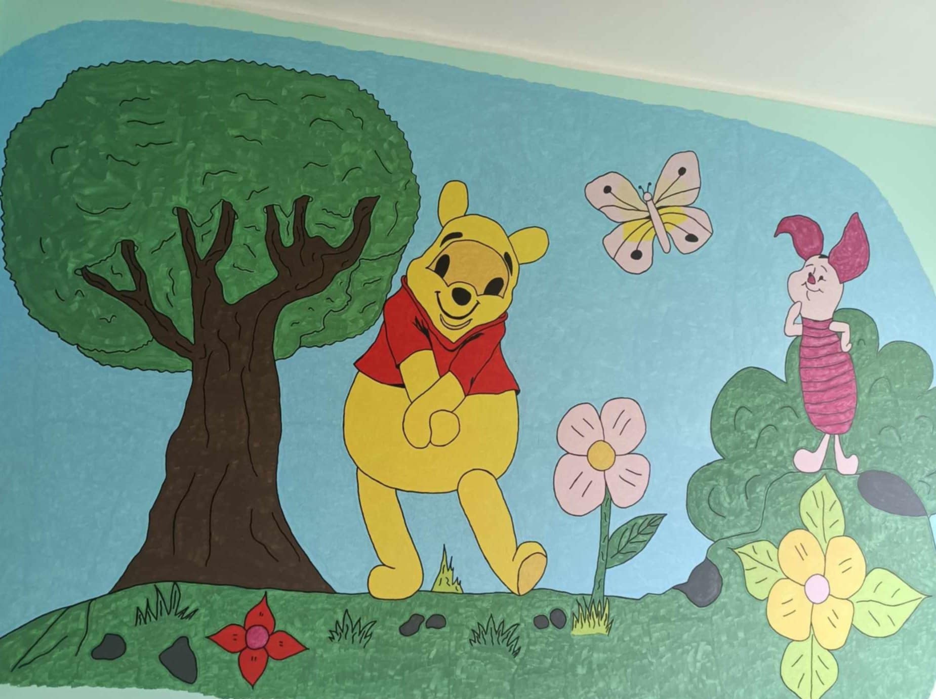 Ręcznie malowana scena z Kubusiem Puchatkiem i Prosiaczkiem na ścianie pokoju dziecięcego, z drzewem, kwiatami i motylem na jasnoniebieskim tle.