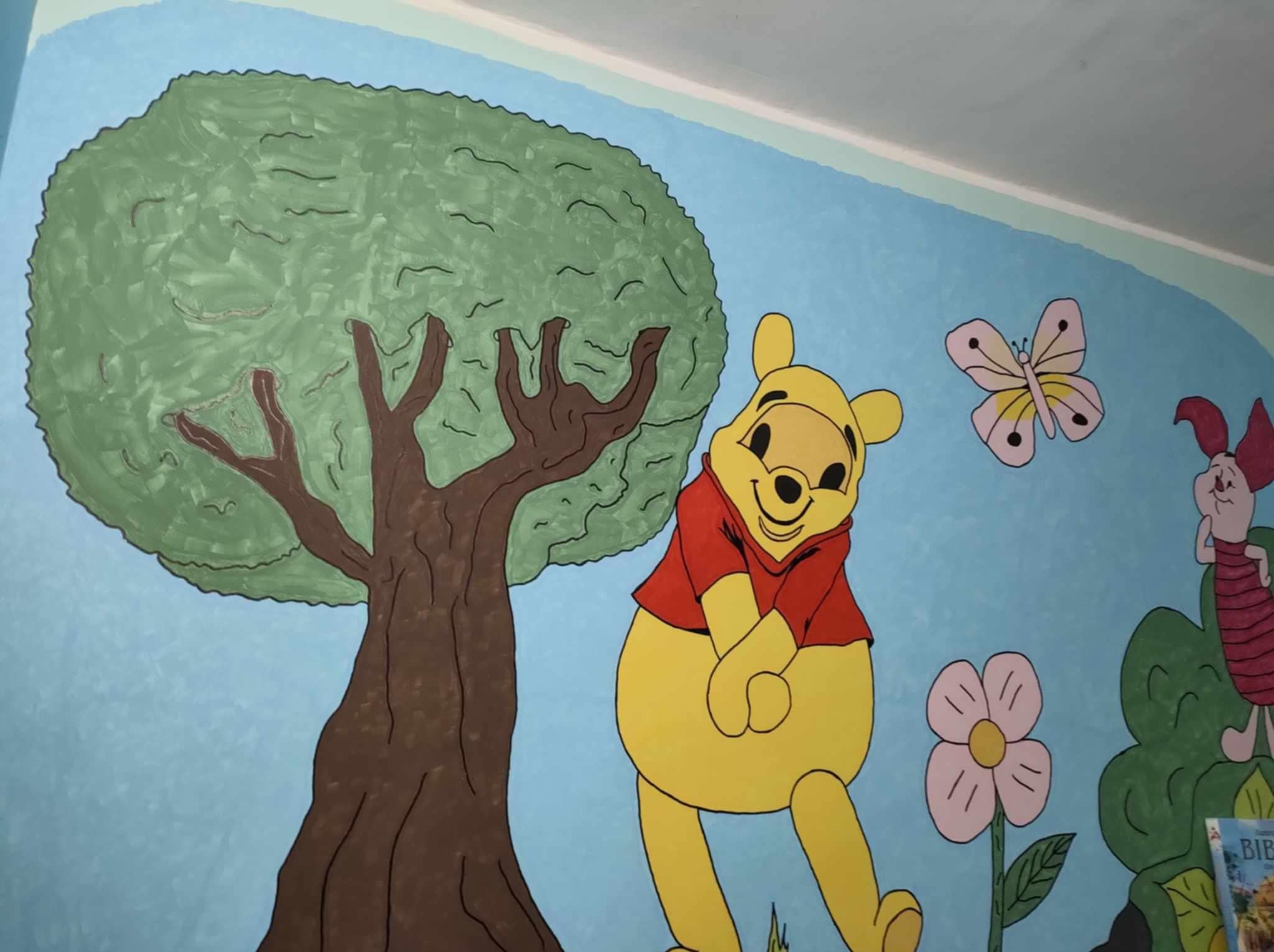Pokój dziecięcy z ręcznie malowaną ścianą: Kubuś Puchatek, drzewo, motyl i Prosiaczek na tle błękitnego nieba. Artystyczne malowanie ścian w pokoju dziecka.