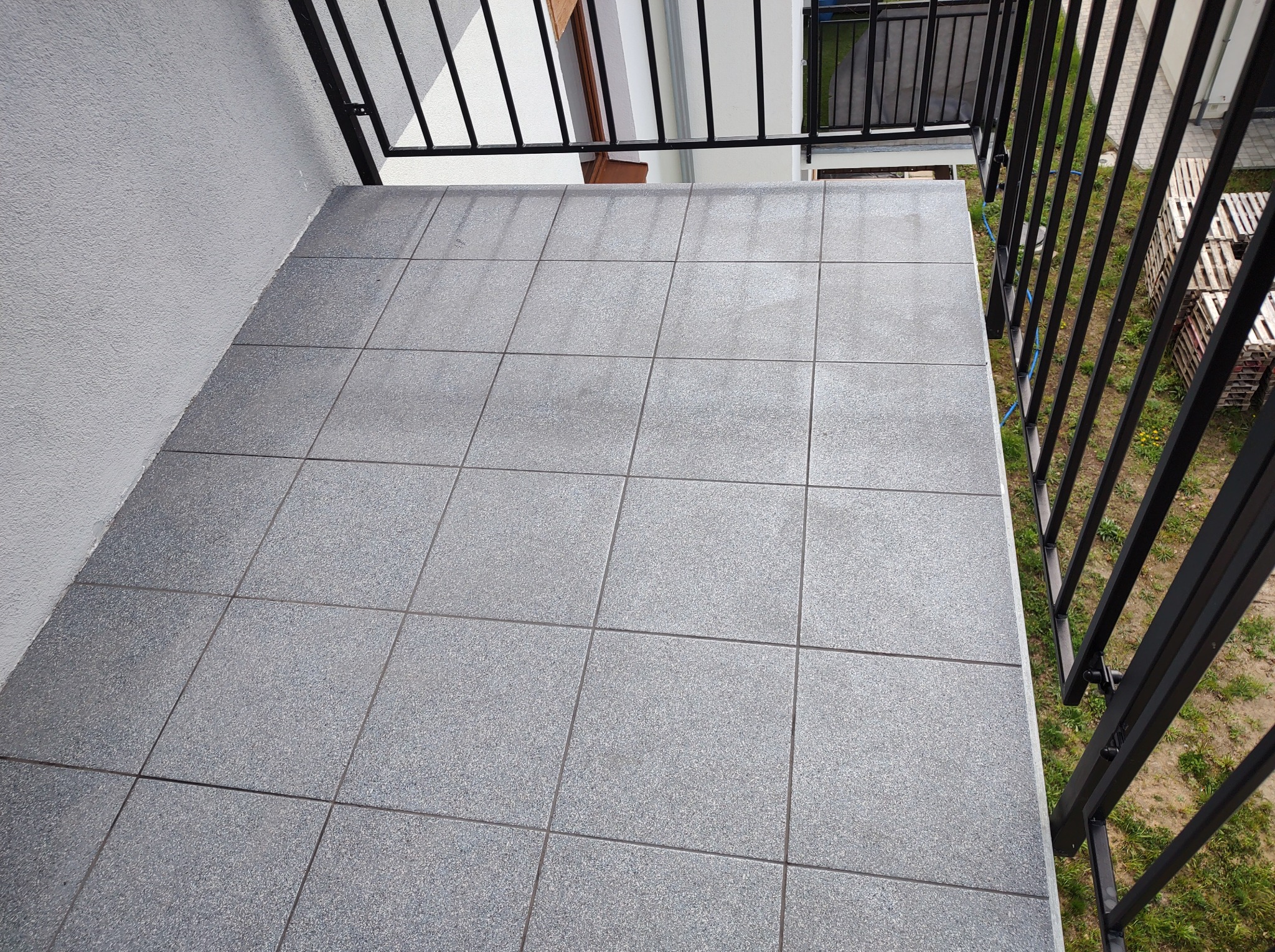 Szara, kwadratowa terakota na balkonie z czarną metalową balustradą. Widok z góry, równomierne oświetlenie, lekko szorstka faktura płytek.