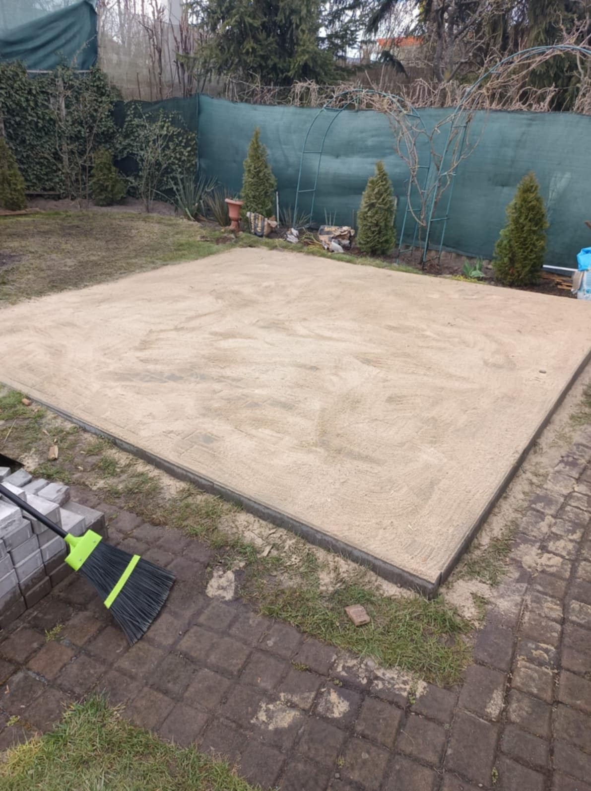 Przygotowany teren pod budowę tarasu z obrzeżami, zasypany piaskiem, z boku leżą kostki brukowe i stoi miotła. Prace ziemne w ogrodzie.