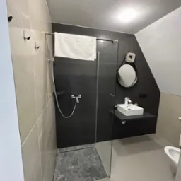 Nowoczesna łazienka z kabiną prysznicową typu walk-in, czarnymi płytkami na ścianie, owalnym lustrem i minimalistyczną umywalką nablatową. Szare płytki podłogowe.