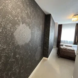 Stylowa ściana z dekoracyjnym wzorem w odcieniach szarości w sypialni. Widoczne dwa łóżka z brązową pościelą i białe ręczniki. Całość dopełnia jasna podłoga.