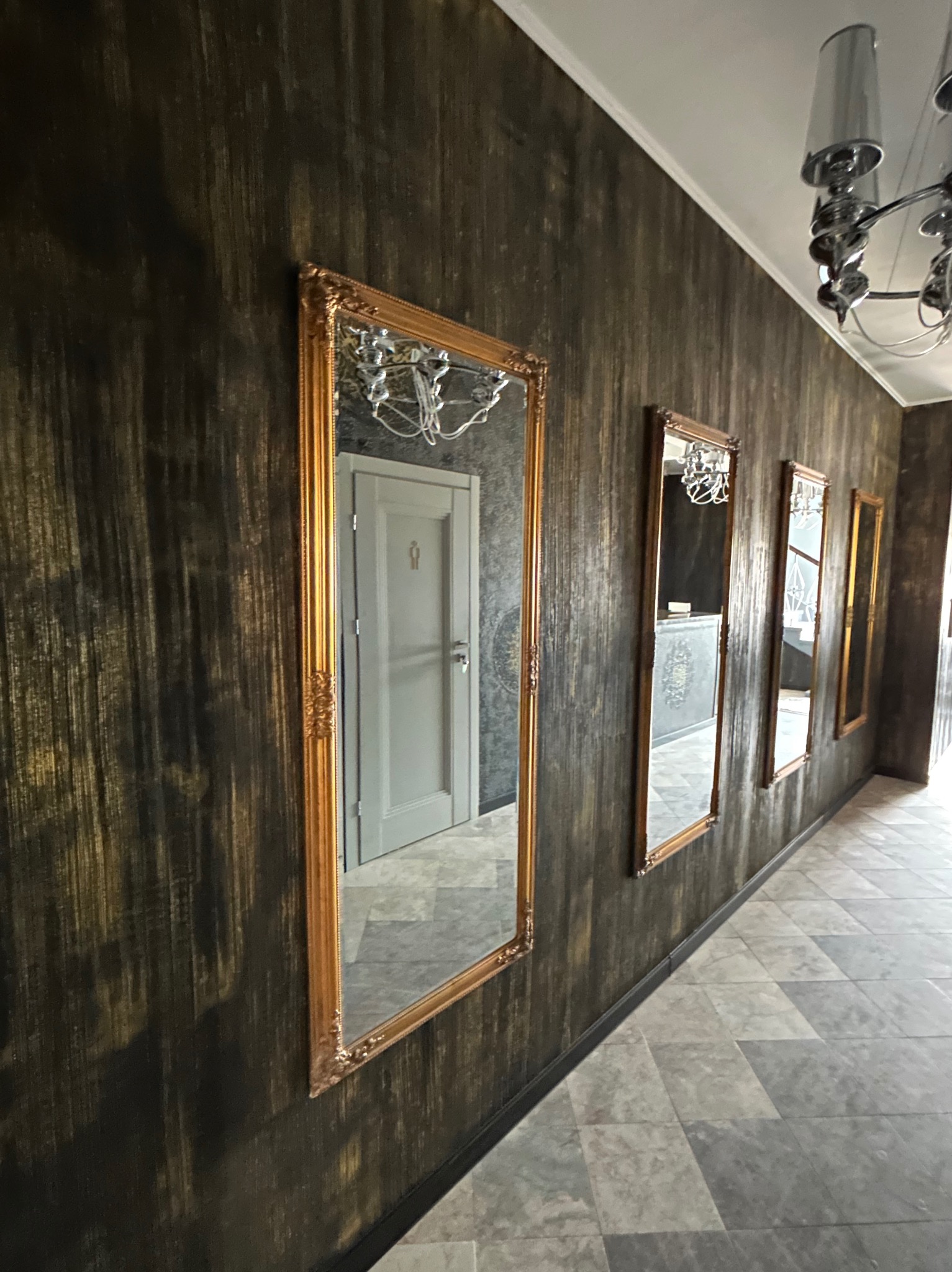 Elegancki korytarz z dekoracyjną ścianą w odcieniach brązu i złota, ozdobiony serią luster w złotych ramach. Widoczna podłoga z szarych płytek i oświetlenie sufitowe.