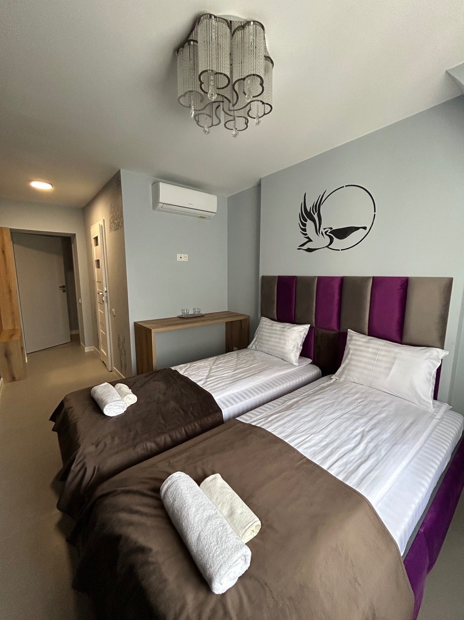 Jasne wnętrze pokoju z dwoma łóżkami, fioletowym wezgłowiem i dekoracją ścienną z motywem ptaka. Widoczny klimatyzator i nowoczesna lampa sufitowa.