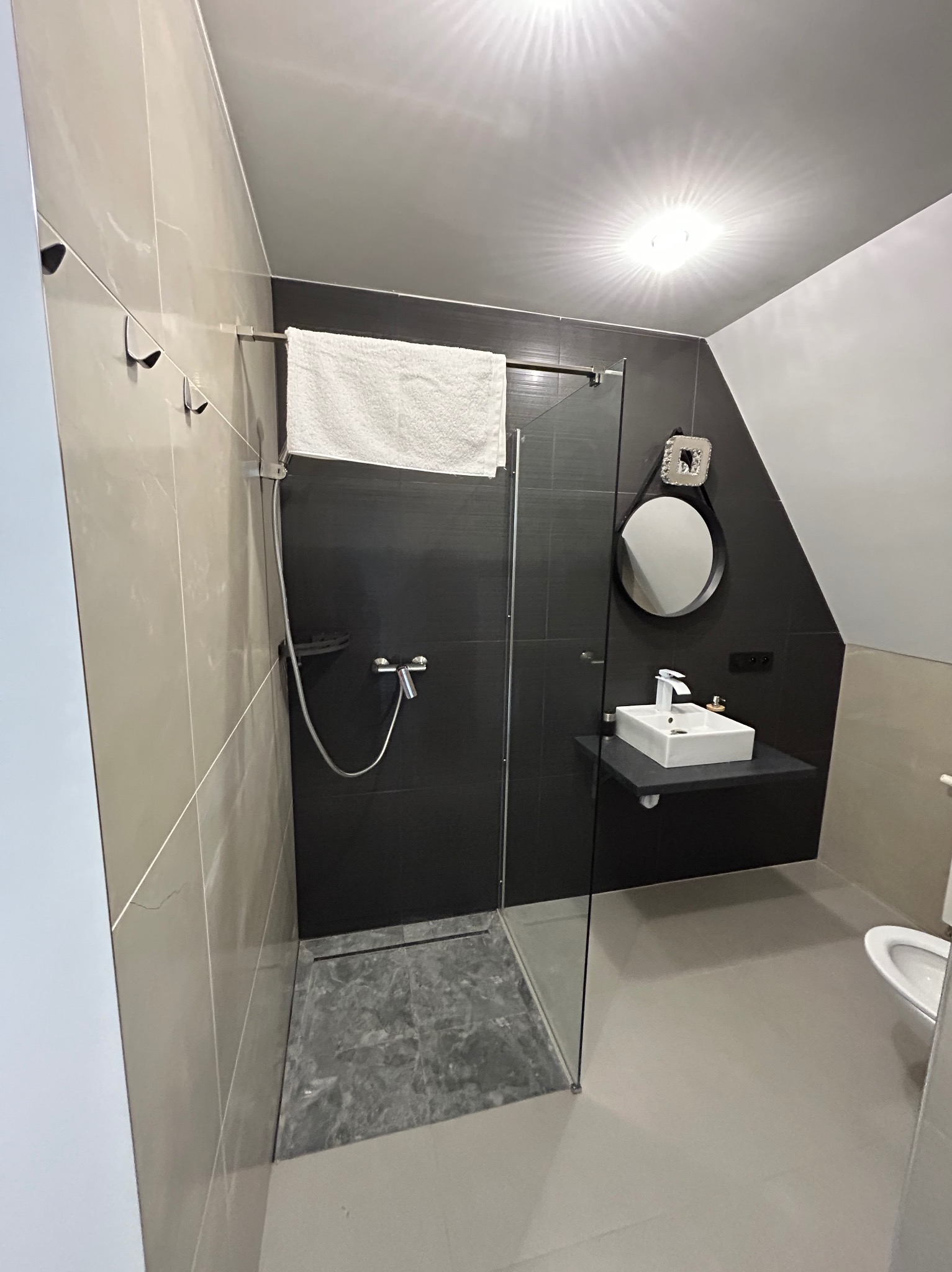 Nowoczesna łazienka z kabiną prysznicową typu walk-in, czarnymi płytkami na ścianie, owalnym lustrem i minimalistyczną umywalką nablatową. Szare płytki podłogowe.